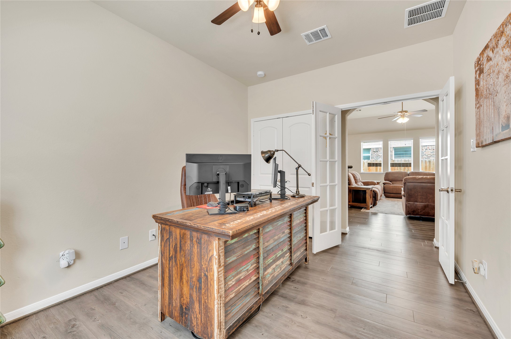Charming home office with rustic desk, ceiling fan, and French doors leading to a cozy living area with ample natural light.
