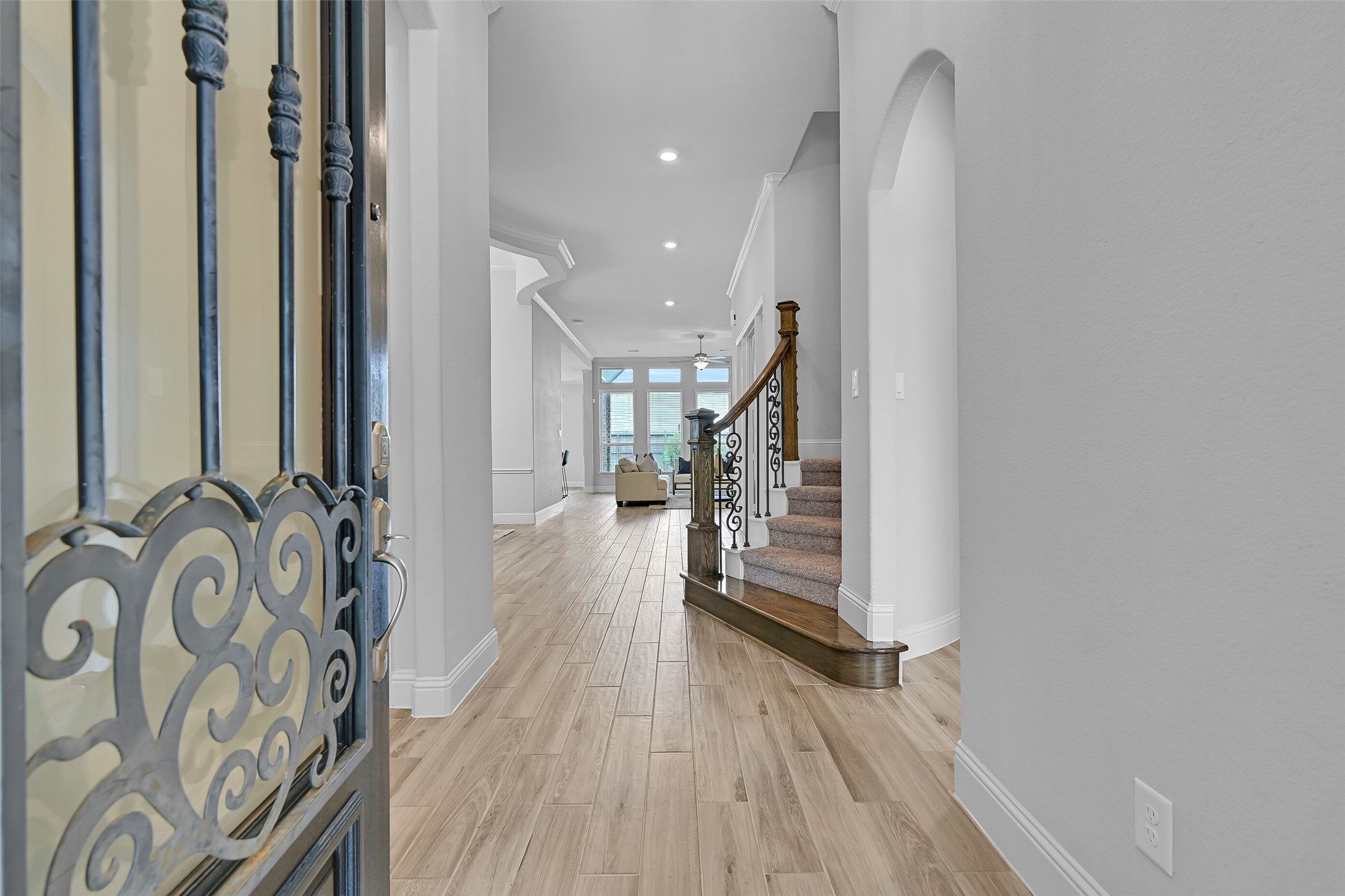 Step inside to a grand foyer featuring wood-look tile flooring, fresh interior paint, crown molding, inset art niches, and a striking split staircase with wood and iron handrails. The open sightline straight through to the family room immediately showcases the home's spacious design.