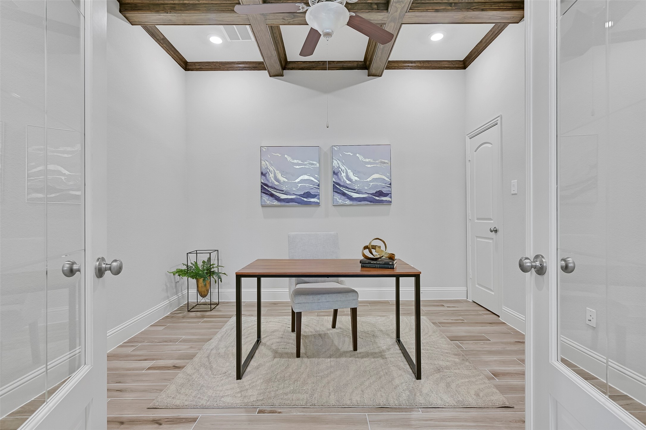 Perfect for working from home, this study features wood-look tile flooring, a coffered ceiling with rich beam detail, recessed lighting, a ceiling fan, French doors with glass inserts, and a convenient closet for additional storage.