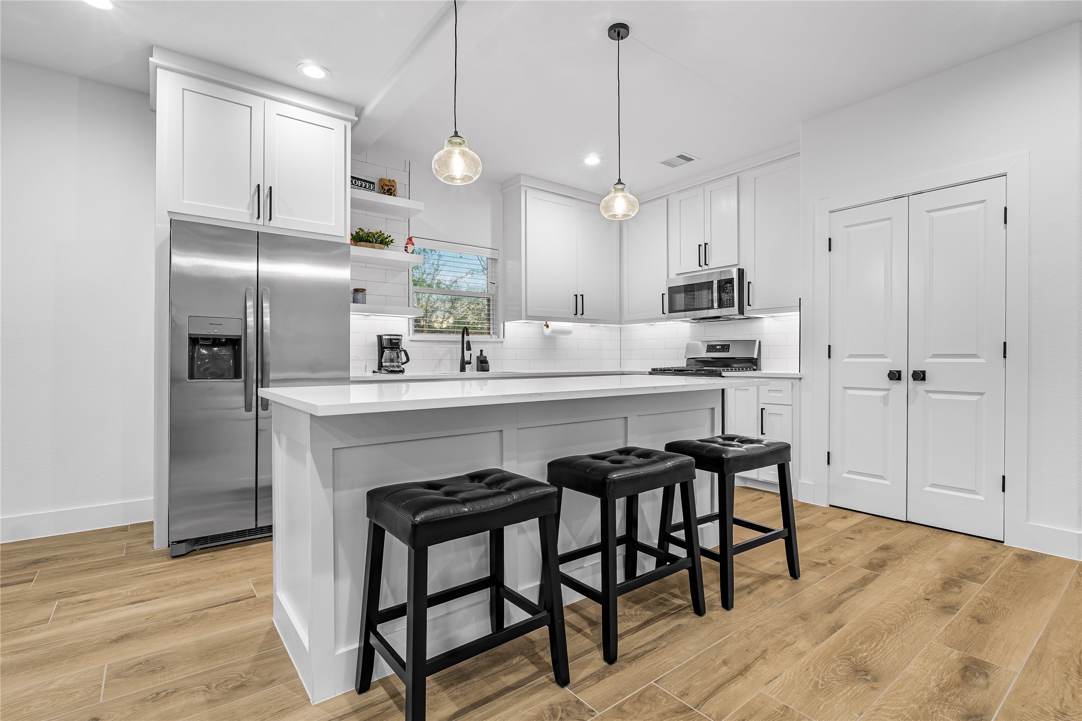Bright and modern kitchen features sleek white cabinetry, a spacious island with seating, stainless steel appliances, and stylish pendant lighting. Actual furniture.