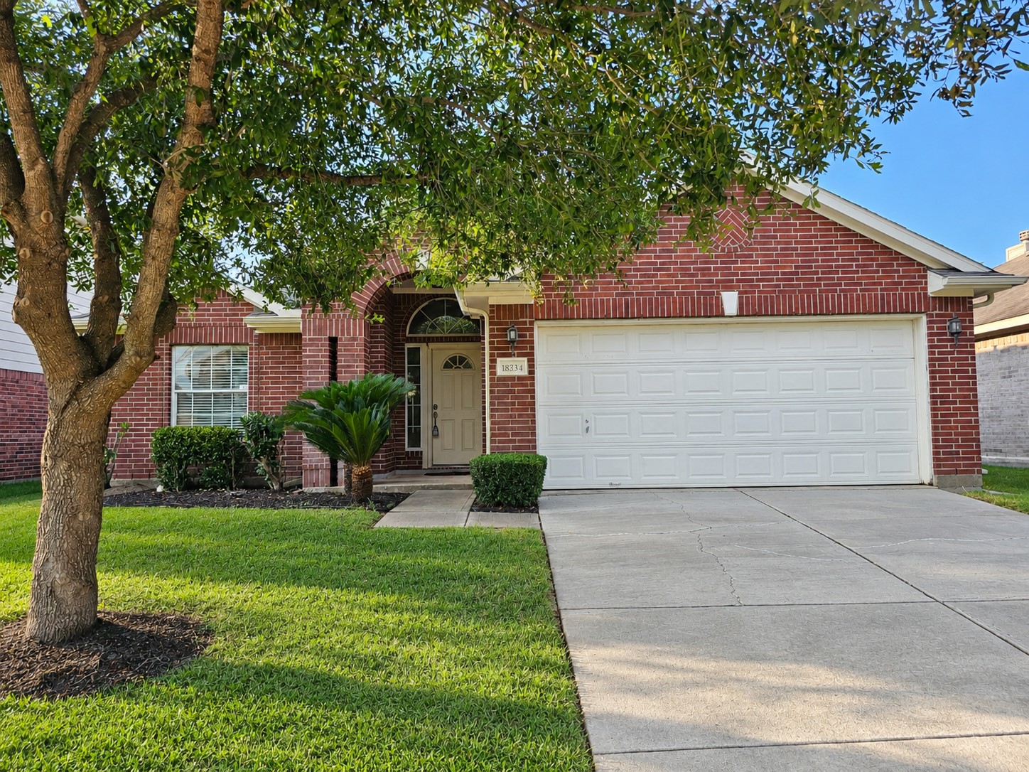 Charming brick home with a two-car garage, nestled under a mature tree, featuring a well-maintained front lawn and inviting entryway.