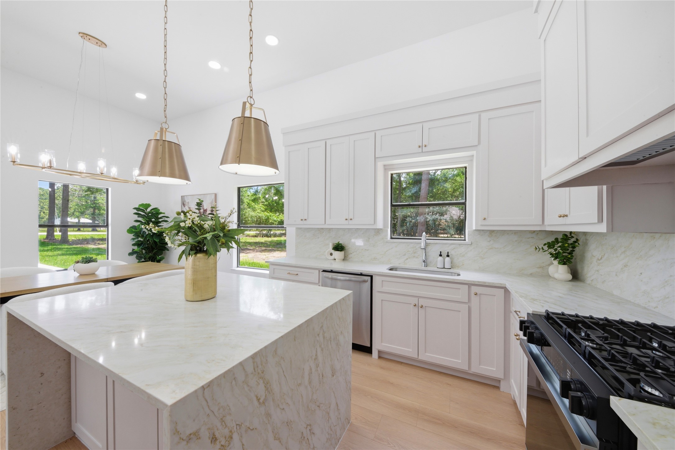 Light-filled kitchen showcasing dual brass pendants over the waterfall quartz island, statement linear chandelier above breakfast nook, walls of windows overlooking wooded acreage, and custom cabinetry with brass hardware.