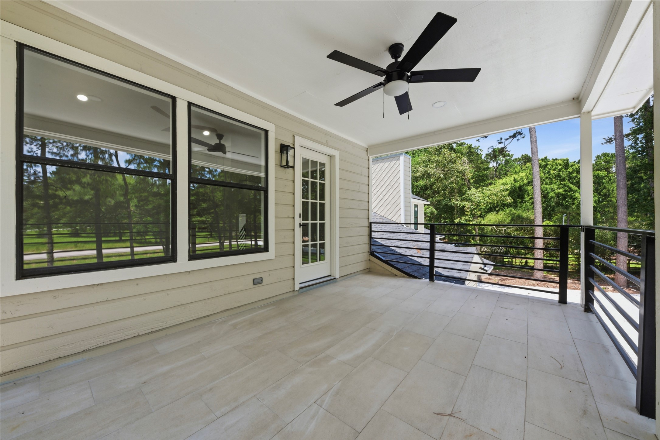 Expansive covered upper balcony showing the rooftop view of the detached 3-car garage structure to the right. French door access from the second primary suite. Horizontal black metal rail with treeline beyond.