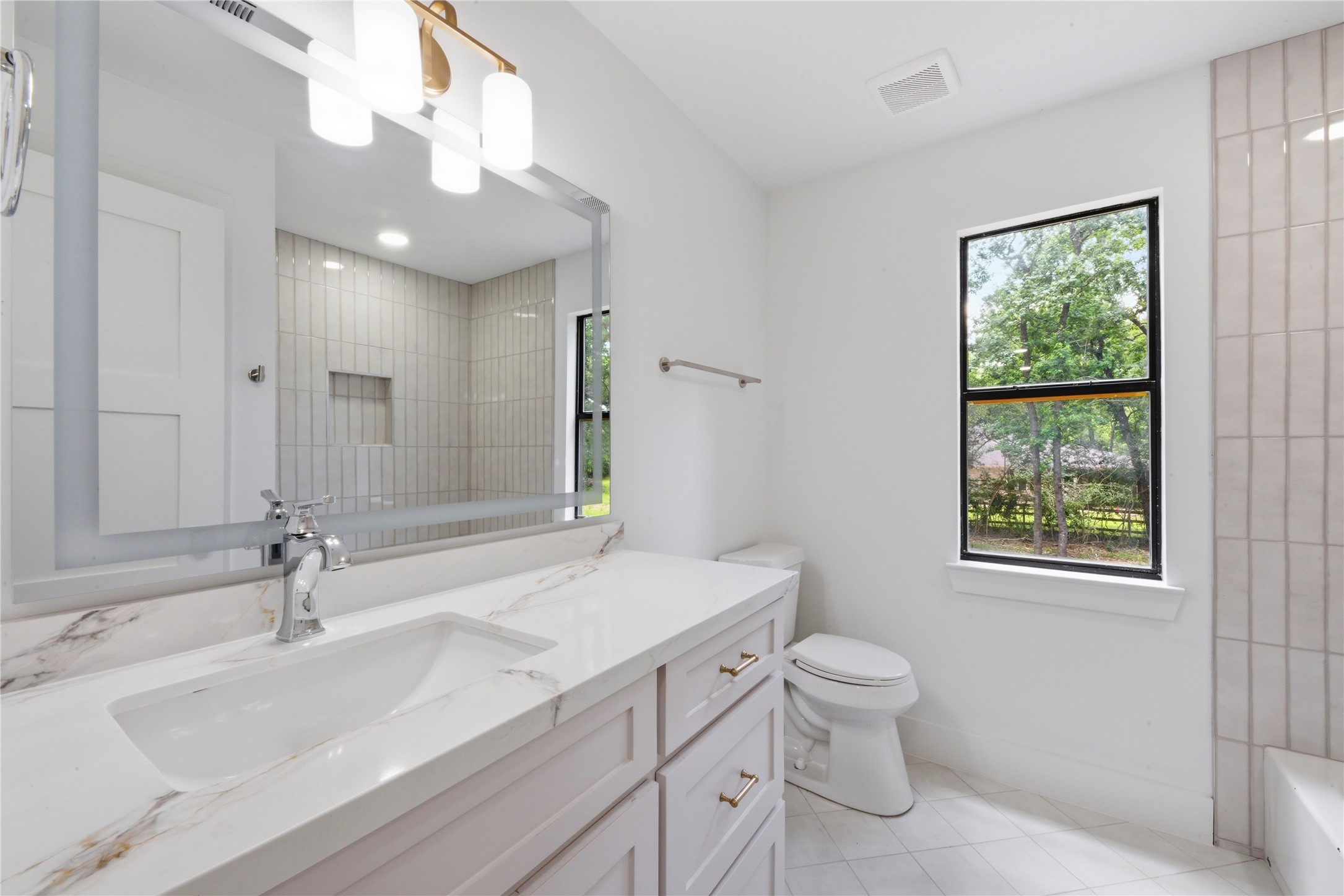 Designer secondary bath featuring quartz counter with marble veining, brass sconces, vertical tile tub/shower surround with niche, and natural light from oversized window. Brass hardware throughout.
