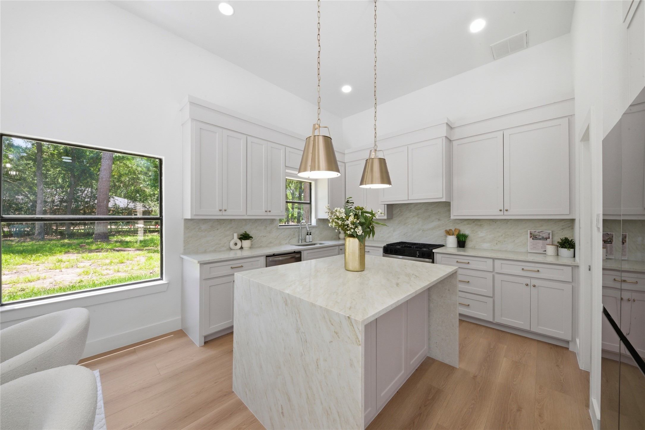 Show-stopping waterfall quartz island anchors this chef's kitchen. Custom cabinetry throughout, brass cone pendants, gas range, marble-look quartz backsplash, and oversized window framing the wooded lot view.
