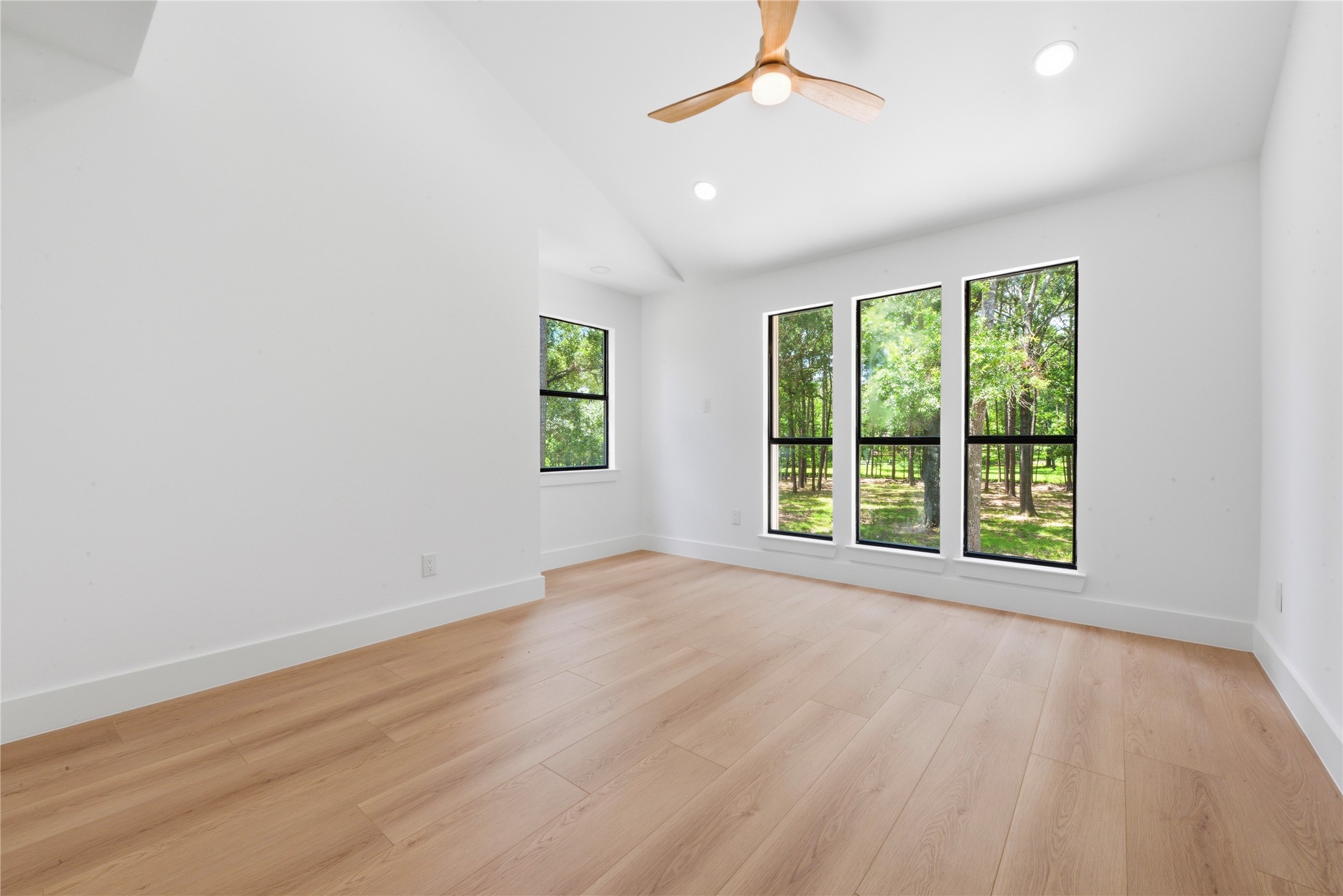 Second upstairs bedroom with vaulted ceiling and designer ceiling fan. Floor-to-ceiling black-frame windows fill the space with natural light and frame the mature tree views.
