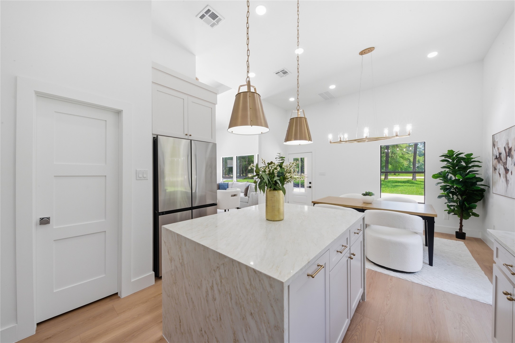 Open-concept kitchen opening to the living and breakfast areas. Soft-close custom cabinetry conceals a walk-in pantry door (left). Refrigerator included. Statement chandeliers and brass pendants illuminate the space.