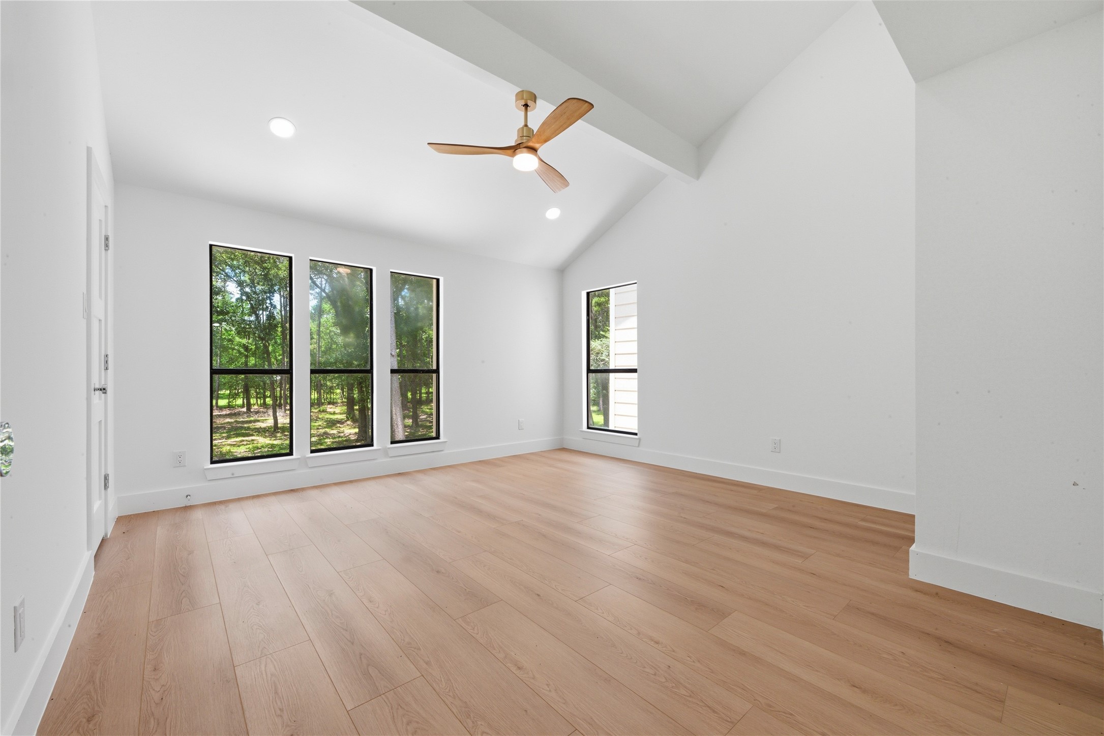 Light-filled secondary bedroom with vaulted ceiling, designer ceiling fan, wide-plank floors, and trio of oversized black-frame windows showcasing wooded acreage views. Generous footprint with closet.