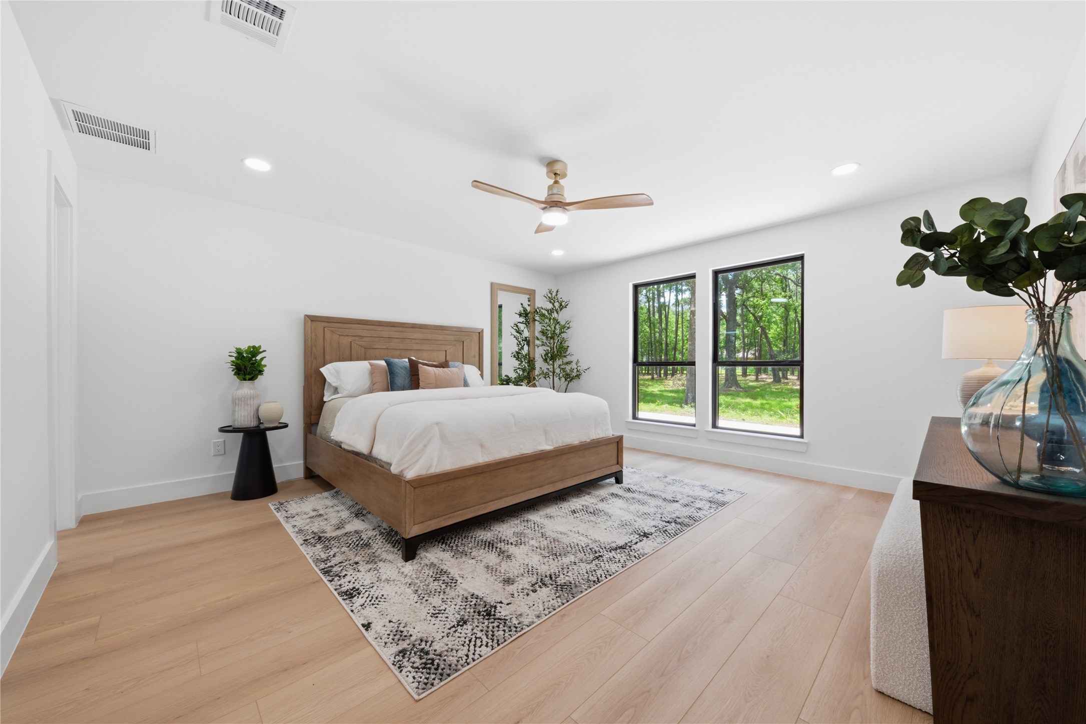 Spacious first-floor primary suite with wide-plank white oak floors, designer ceiling fan, oversized windows framing wooded backyard views, and direct ensuite access. Light-filled and serene retreat.