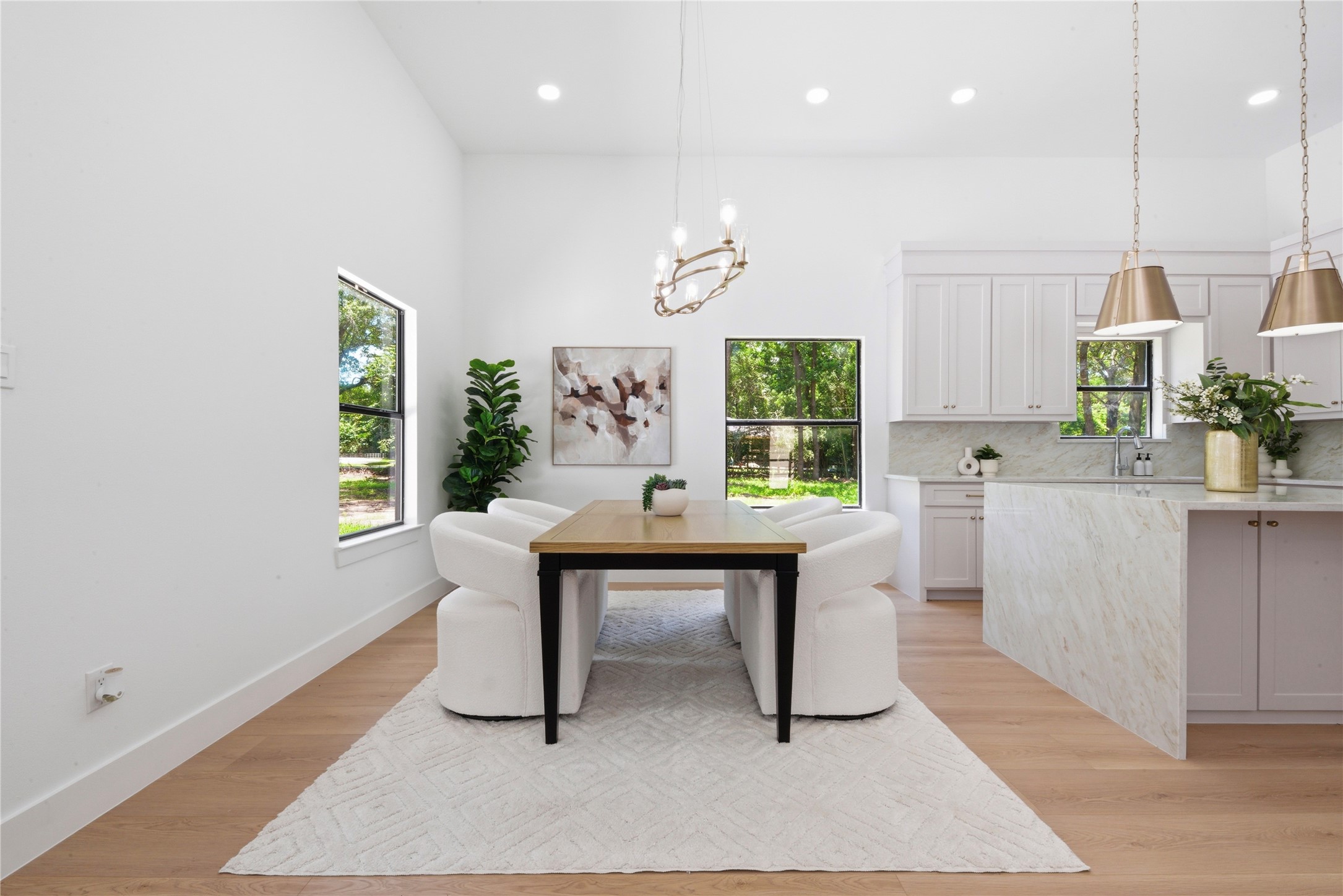 Dining space flowing into the chef's kitchen featuring waterfall quartz island and brass pendant lighting. Vaulted ceilings, designer chandelier, and oversized windows frame views of the wooded acreage.