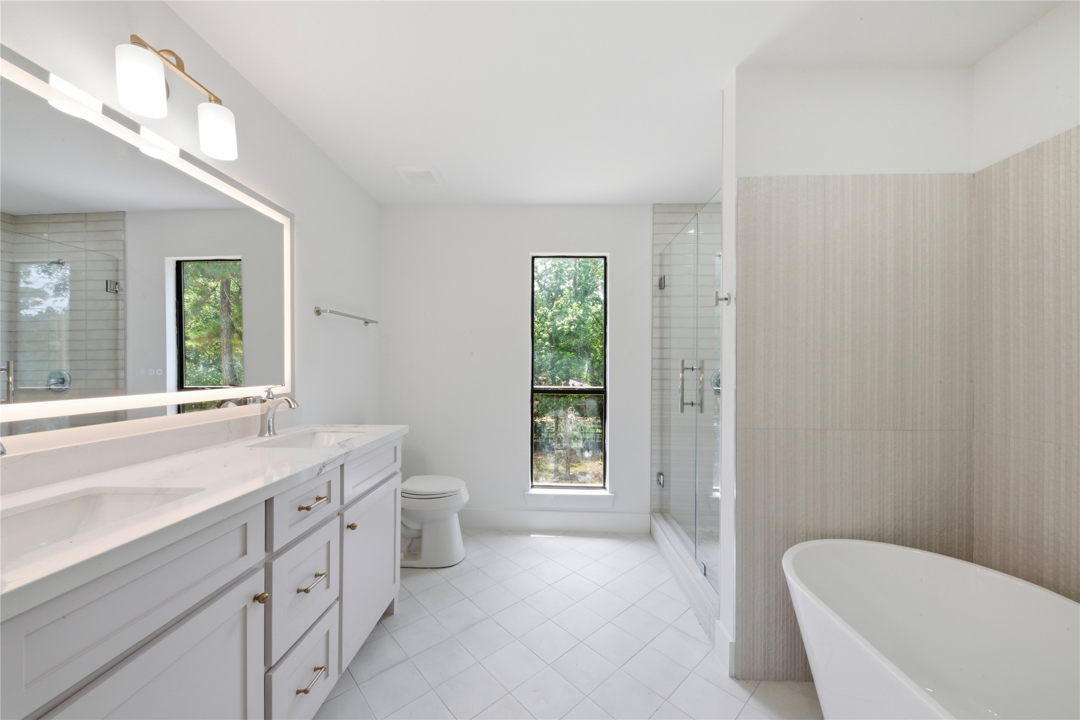 Luxurious fourth-floor ensuite bath serving the second primary suite. Features dual quartz vanity, LED-backlit mirror, brass fixtures, freestanding soaking tub, frameless glass shower with vertical tile, and serene wooded views.