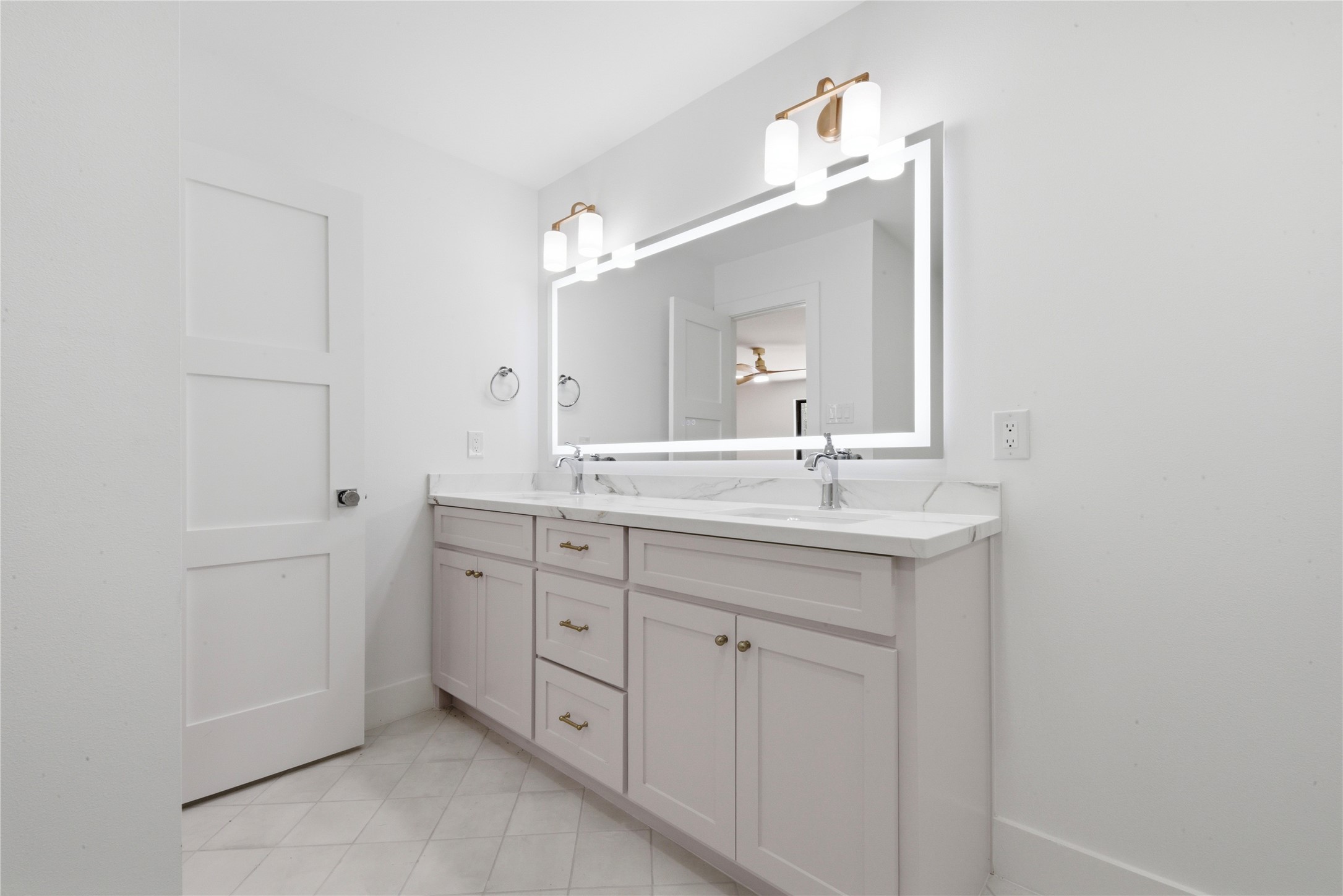 Designer secondary bath featuring dual sinks with quartz waterfall counter, LED-backlit vanity mirror, brass sconces, custom shaker cabinetry with brass hardware, and tile flooring. Shared by the two upstairs secondary bedrooms.