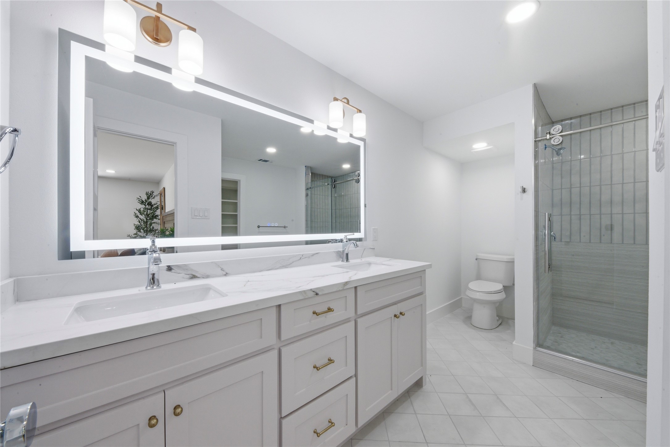Spa-style primary bath featuring dual vanity with quartz counters, LED-backlit designer mirror, brass sconces, large frameless walk-in shower with vertical tile, and separate water closet. Marble-look tile flooring.