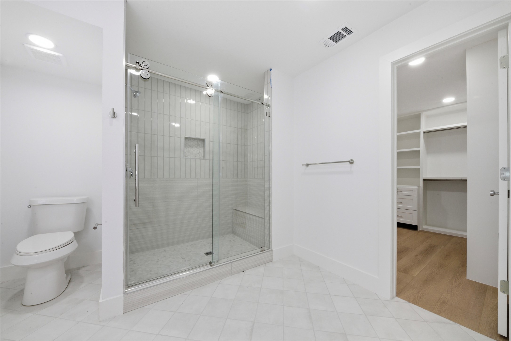 Oversized frameless glass walk-in shower with vertical tile, built-in bench, and shampoo niche. Doorway opens directly to the custom-built walk-in closet, a true ensuite layout.