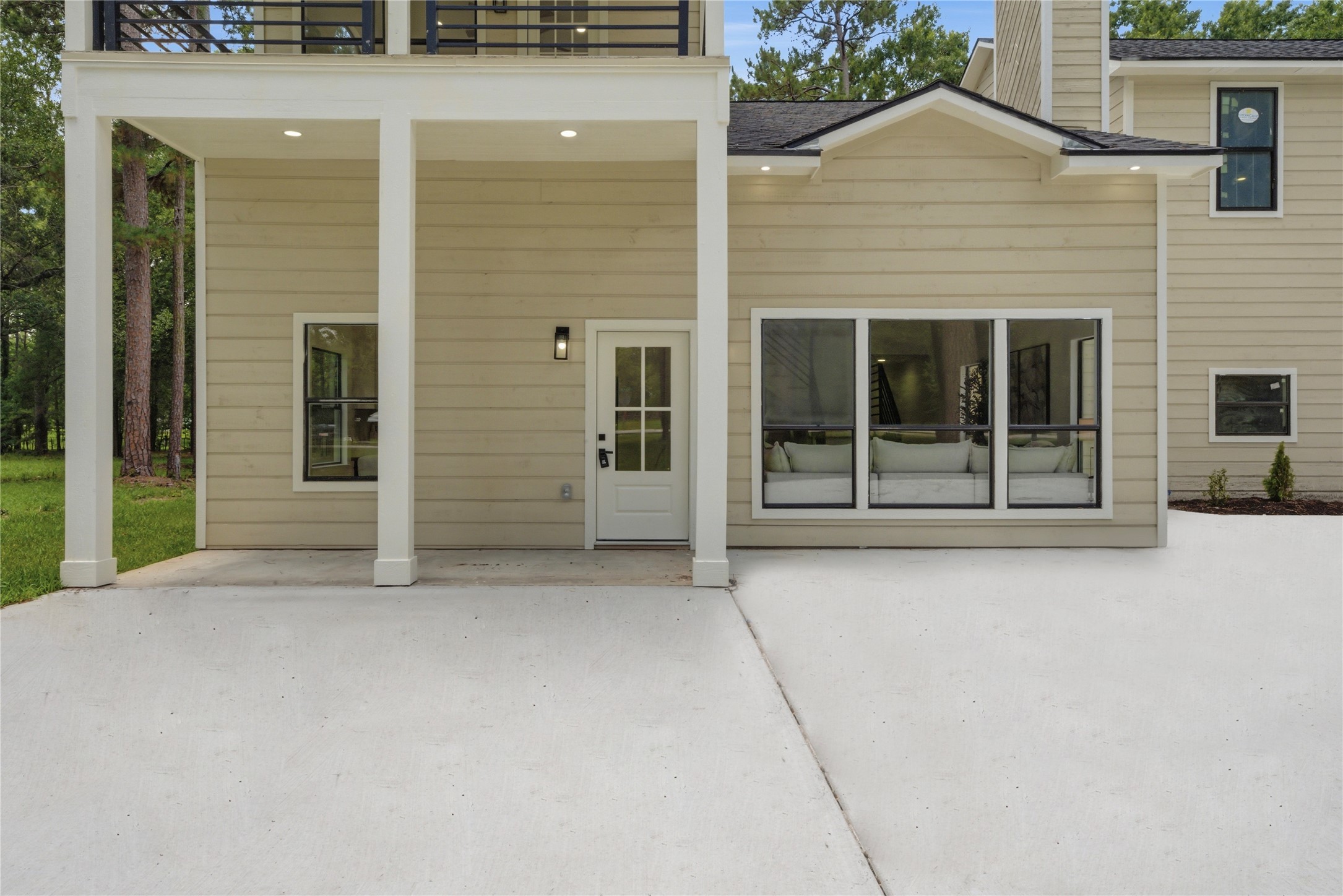 Striking modern farmhouse facade featuring covered upper balcony, double-stack design, board-and-batten siding, and oversized picture windows showcasing the open-concept interior. Pristine concrete drive and walkway.