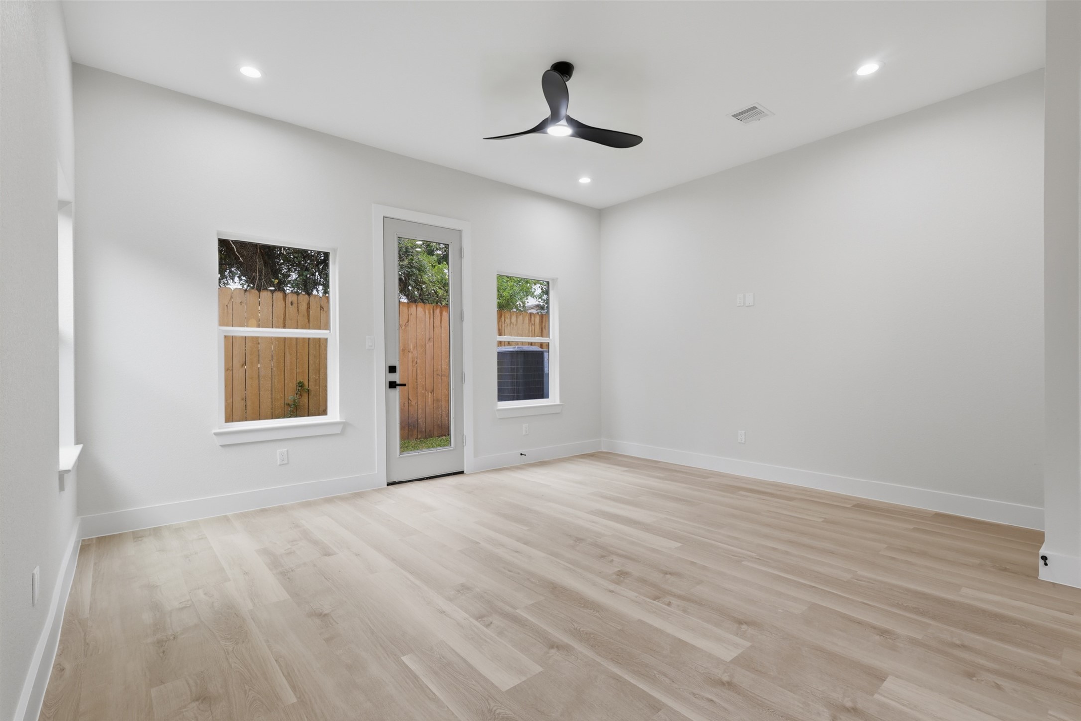 Spacious and bright room featuring sleek LVP floors, modern recessed lighting, and a ceiling fan, with large windows and a glass door leading to a fenced backyard.
