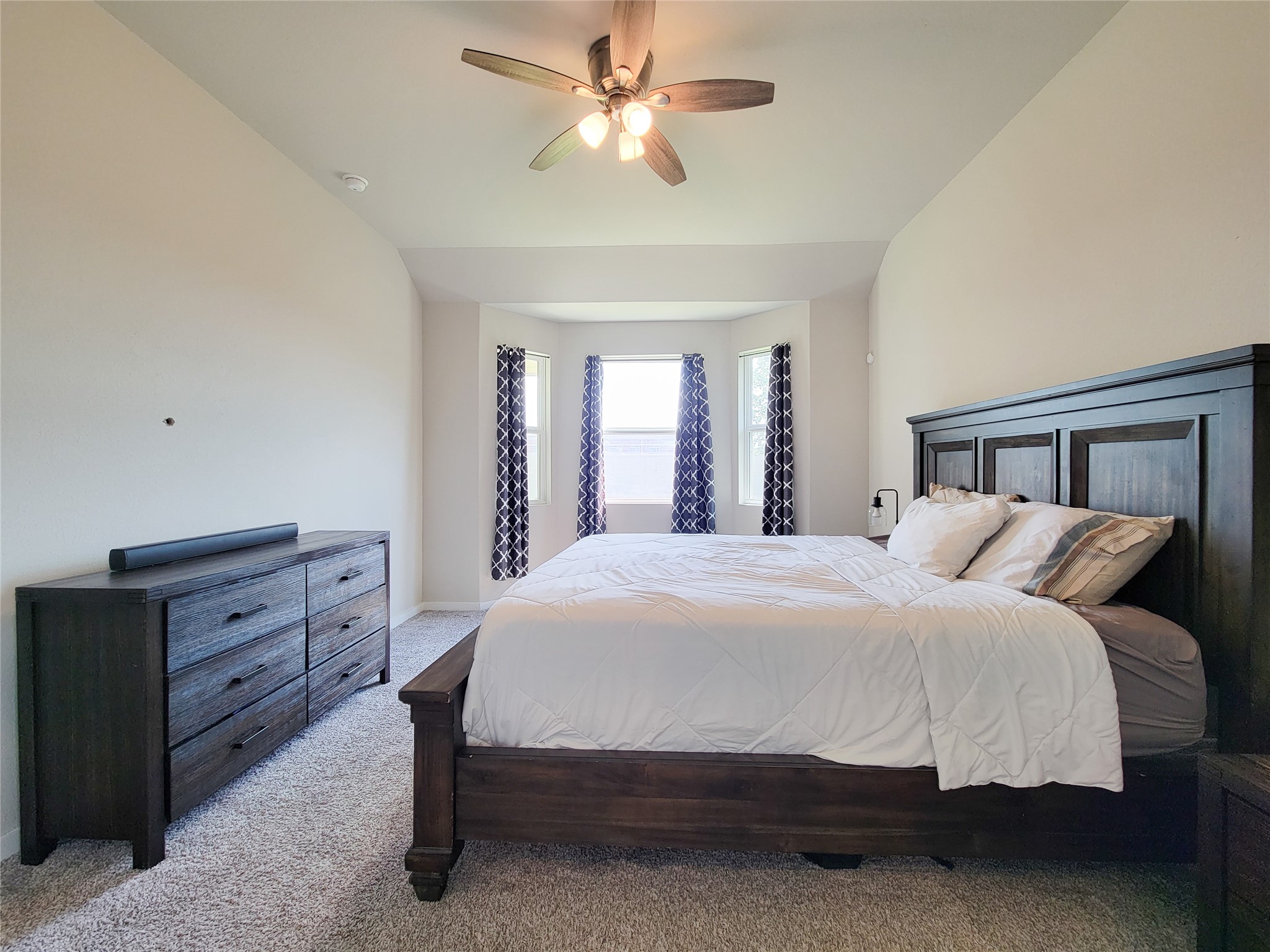Spacious bedroom with a cozy bay window, elegant dark wood furniture, and soft carpeting, complemented by a modern ceiling fan for comfort.