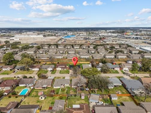 This aerial photo showcases a residential neighborhood with neatly arranged houses and spacious yards. A red marker highlights a specific property. The area appears well-maintained, with some homes featuring pools. In the background, there are commercial and industrial buildings, indicating proximity to urban amenities.