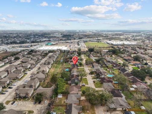 This aerial photo showcases a suburban neighborhood with rows of houses, tree-lined streets, and nearby commercial areas. The area is marked by a prominent pin, indicating a specific property of interest. The surroundings include ample green spaces and easy access to amenities.