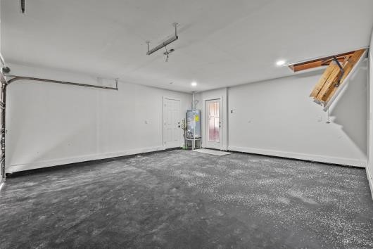 This photo shows a spacious, clean garage with a concrete floor, white walls, and ceiling. It features a water heater, access to the attic via a pull-down ladder, and a door leading outside. The space is well-lit with overhead lighting.