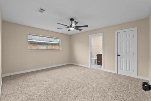 This spacious primary bedroom features neutral-painted walls and carpet, a ceiling fan for comfort, and ample natural light from a large window. It includes an en-suite bathroom and a  walk in closet for convenience.