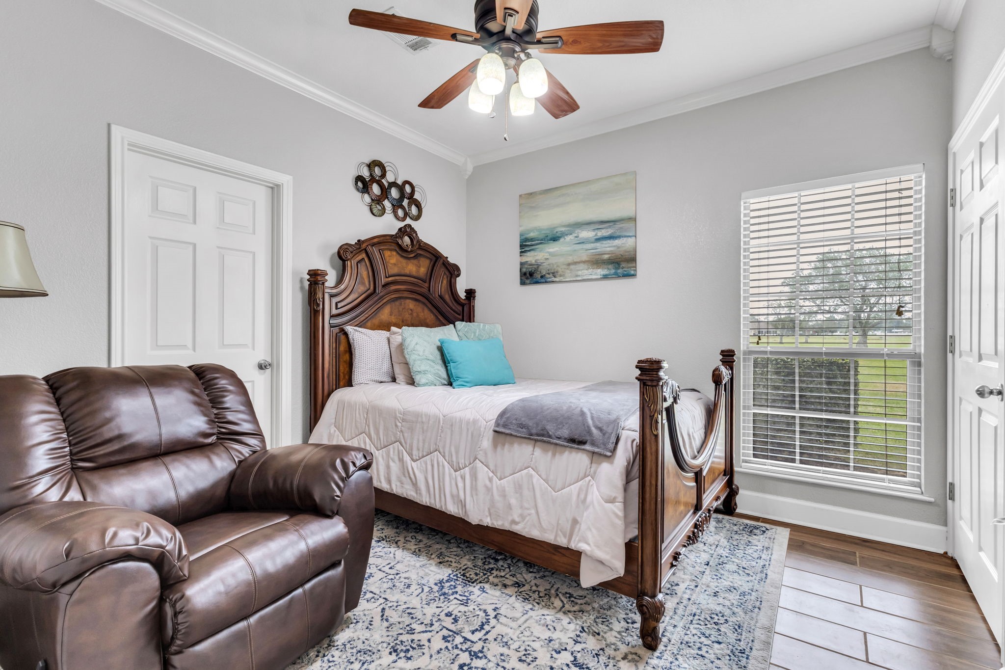 Another view of guest bedroom one, featuring wood-look flooring, crown molding, a ceiling fan, and a large window that brings in natural light. The room offers a comfortable layout with ample closet space.