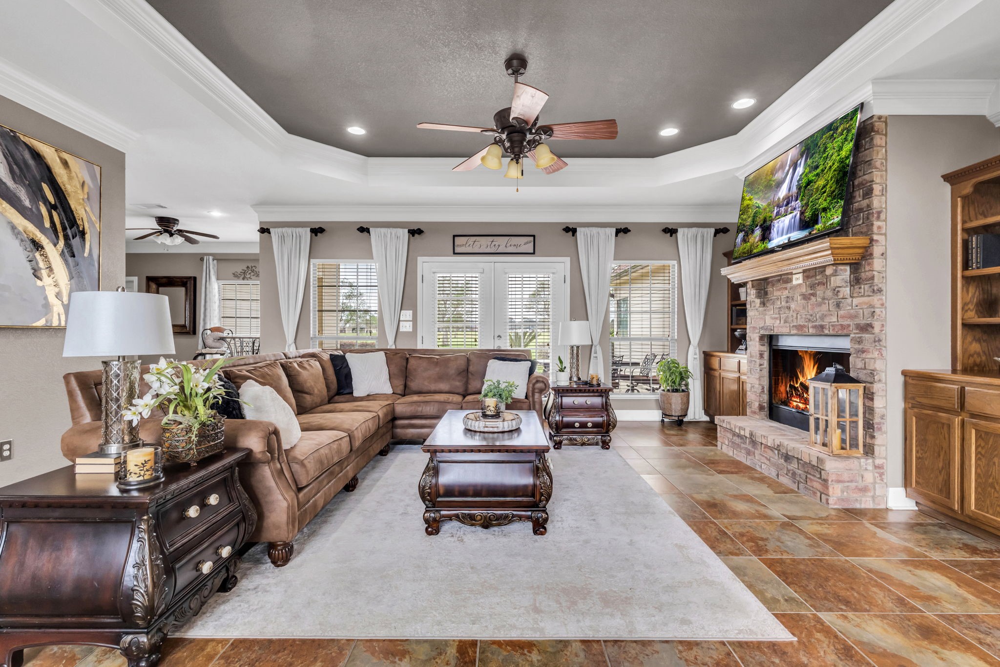 Another view of the spacious living room showcasing the floor-to-ceiling brick fireplace, custom built-in cabinetry, and tray ceiling with crown molding. French doors and surrounding windows offer plenty of natural light and views of the backyard.