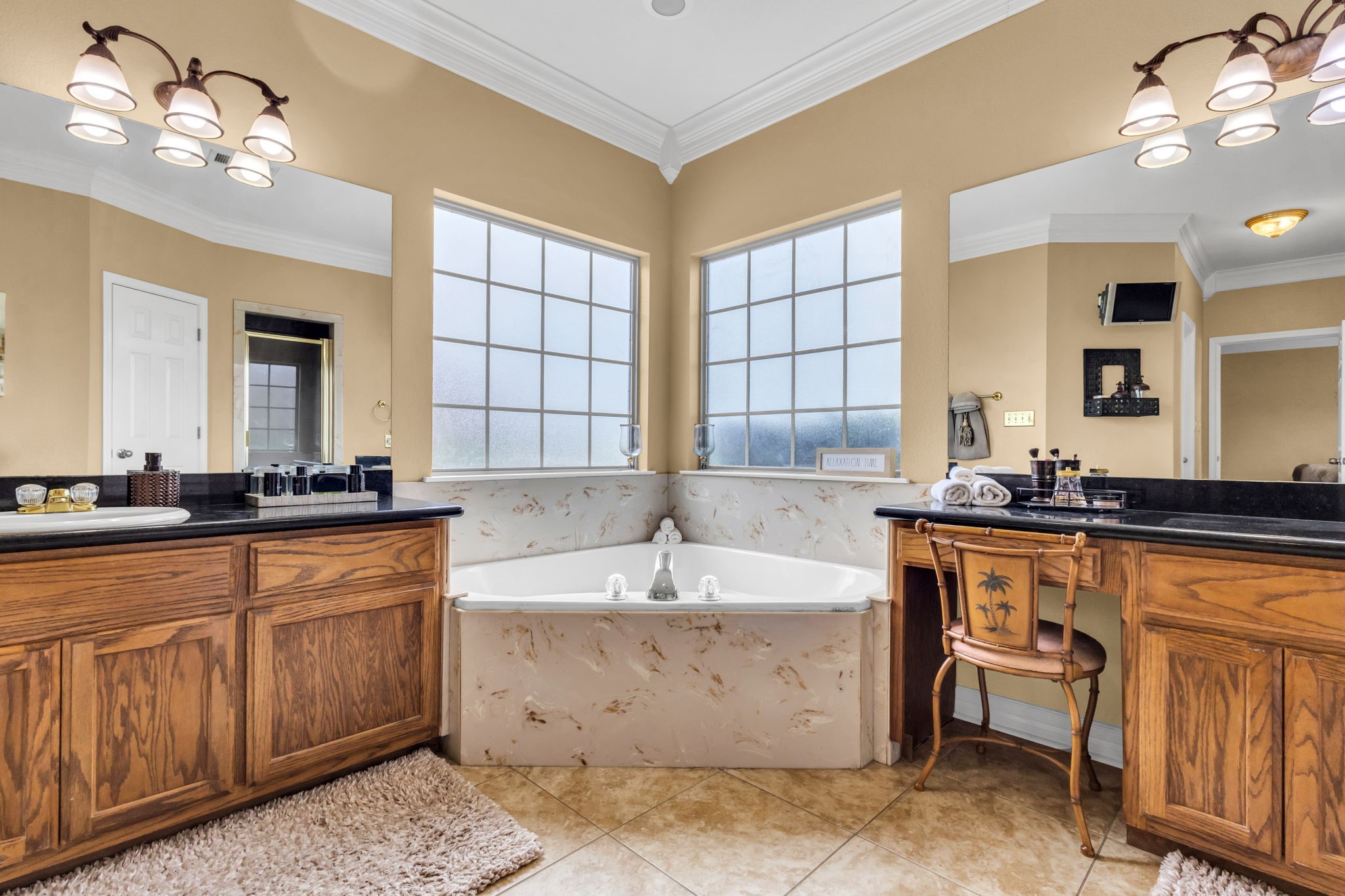 Another view of the spacious primary bathroom highlights the corner soaking tub beneath two large windows, separate vanities with dark countertops, and generous cabinet storage.