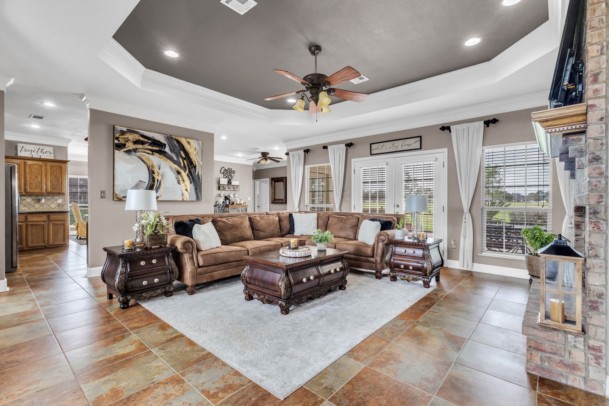 Spacious living room featuring a tray ceiling with crown molding, recessed lighting, tile flooring, and a brick fireplace. Large windows and French doors provide natural light, with an open layout connecting to the kitchen and breakfast area.