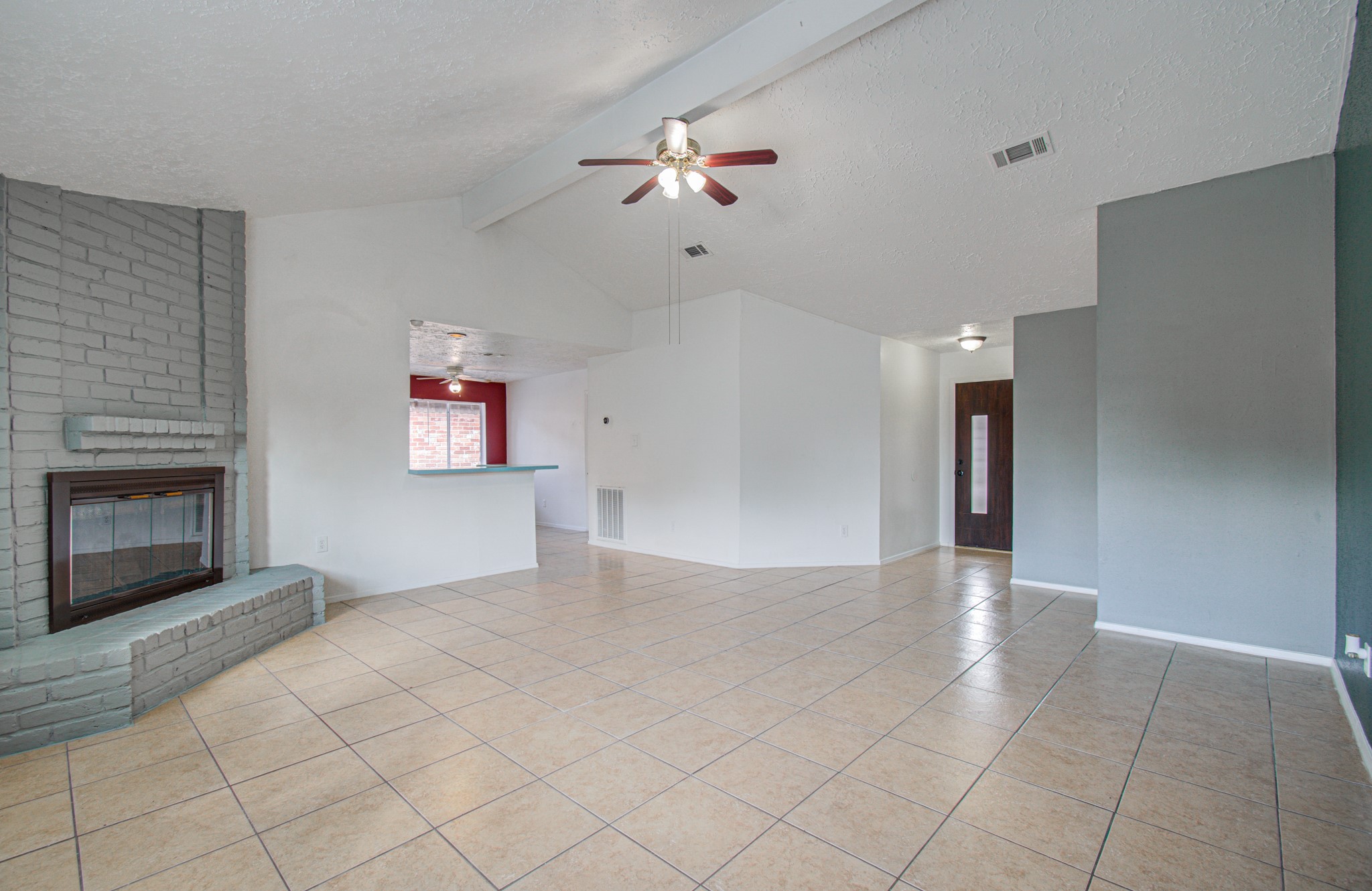 Spacious living area with high ceilings, featuring a cozy brick fireplace and modern ceiling fan, complemented by neutral tile flooring and a pass-through to the kitchen.