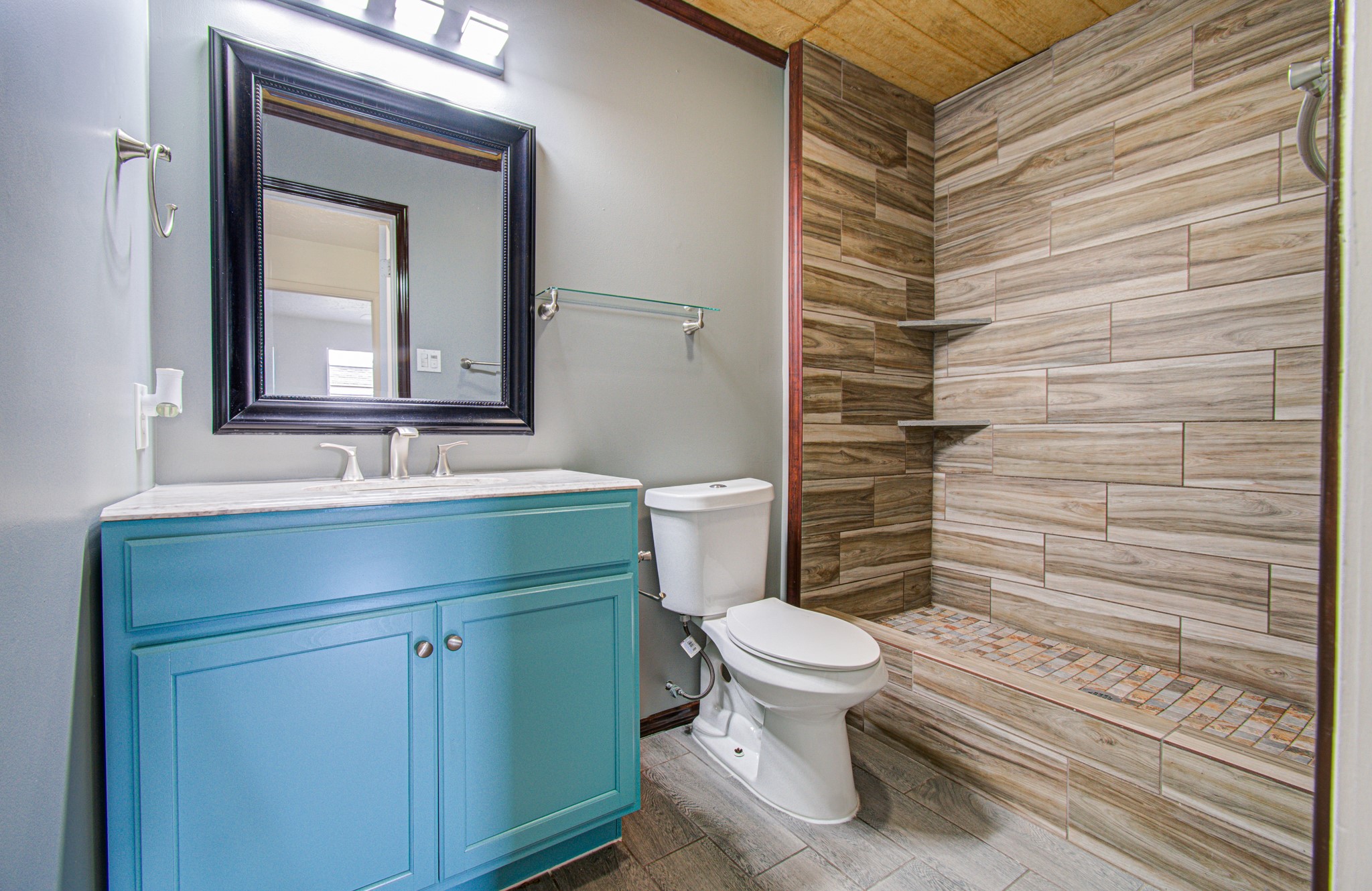 Elegant bathroom featuring a striking wood-look tile shower, sleek blue vanity with modern fixtures, and a large framed mirror.