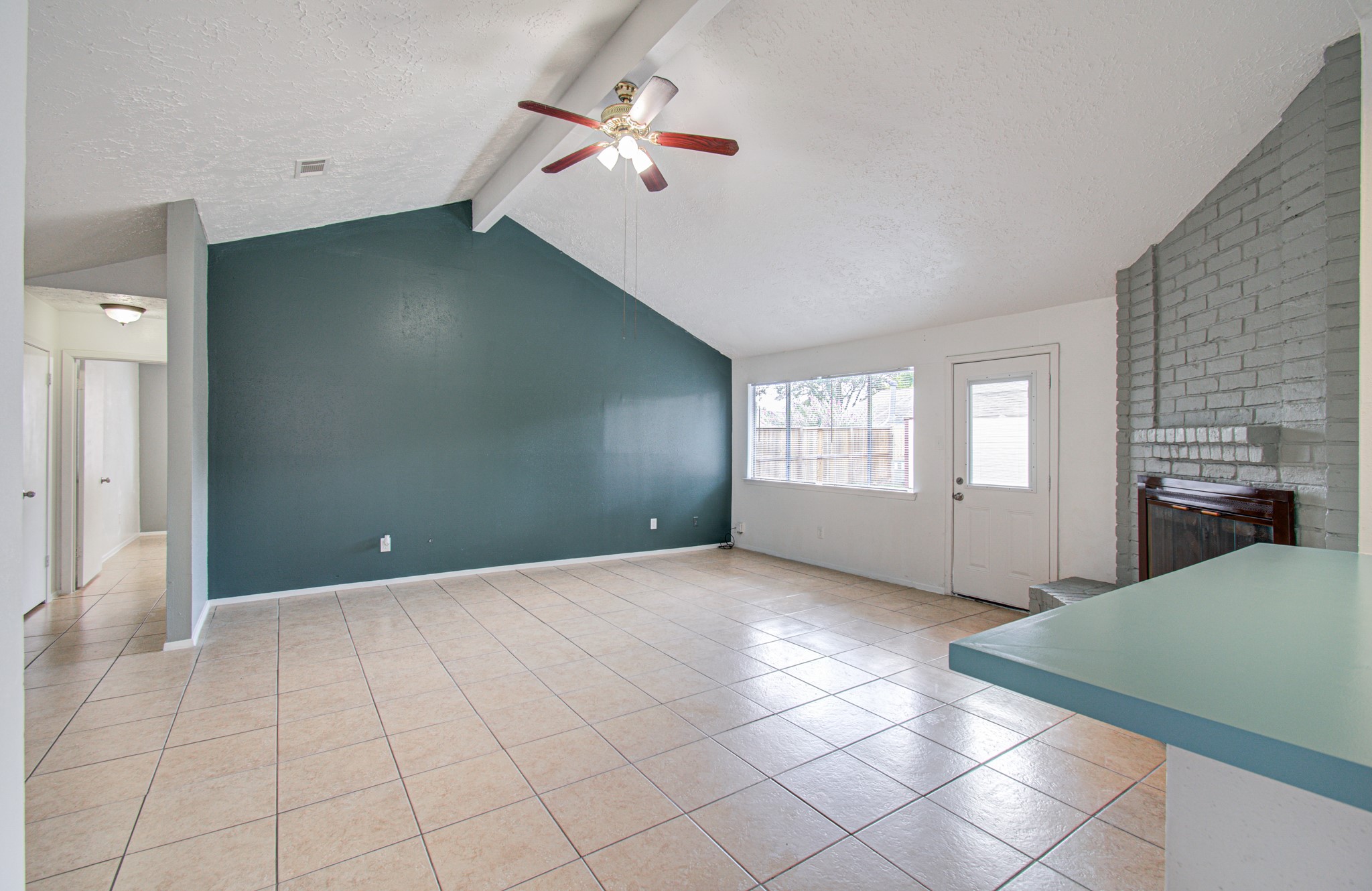 Spacious living area with high ceilings, a ceiling fan, and abundant natural light from large windows. The room features tile flooring, a cozy brick fireplace, and a vibrant accent wall.