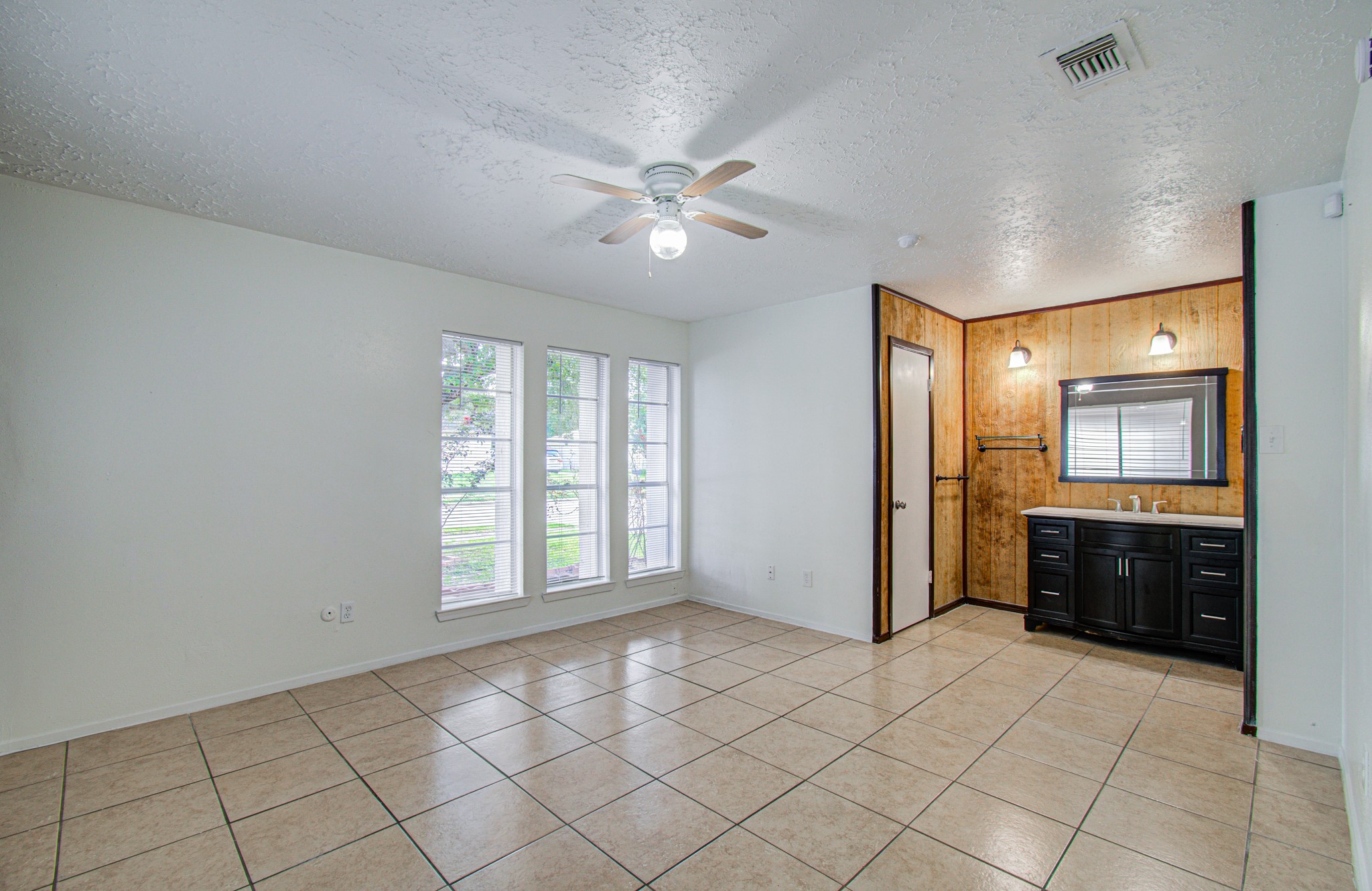 Bright living space with large windows, tiled floors, and a ceiling fan, featuring a cozy corner with a modern vanity and wood accent wall. Bright living space with large windows, tiled floors, and a ceiling fan, featuring a cozy corner with a modern vanity and wood accent wall.