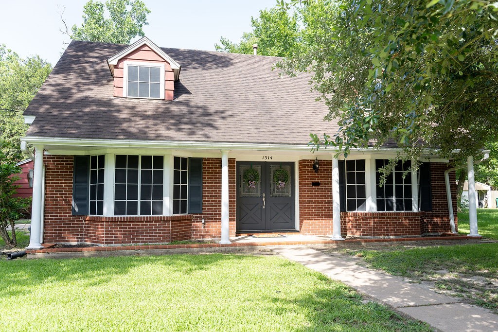 Charming two-story brick home with a gabled roof and dormer window. Features a welcoming front porch, large windows, and a well-maintained lawn, framed by mature trees.