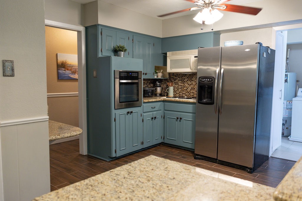 This kitchen features stylish blue cabinets, stainless steel appliances, and granite countertops. The mosaic tile backsplash adds a modern touch, and the ceiling fan provides comfort. Adjacent areas offer additional space and functionality.