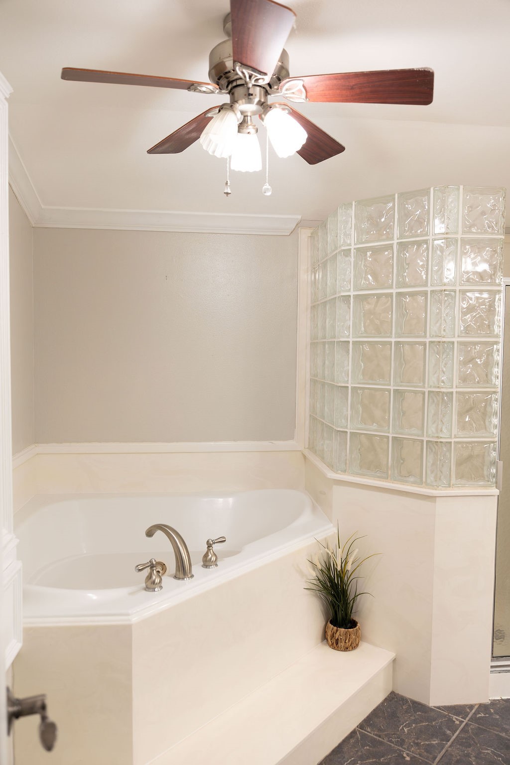 This bathroom features a spacious corner bathtub with modern fixtures, surrounded by elegant glass block walls. A ceiling fan with lights adds comfort, while a small plant provides a touch of greenery.
