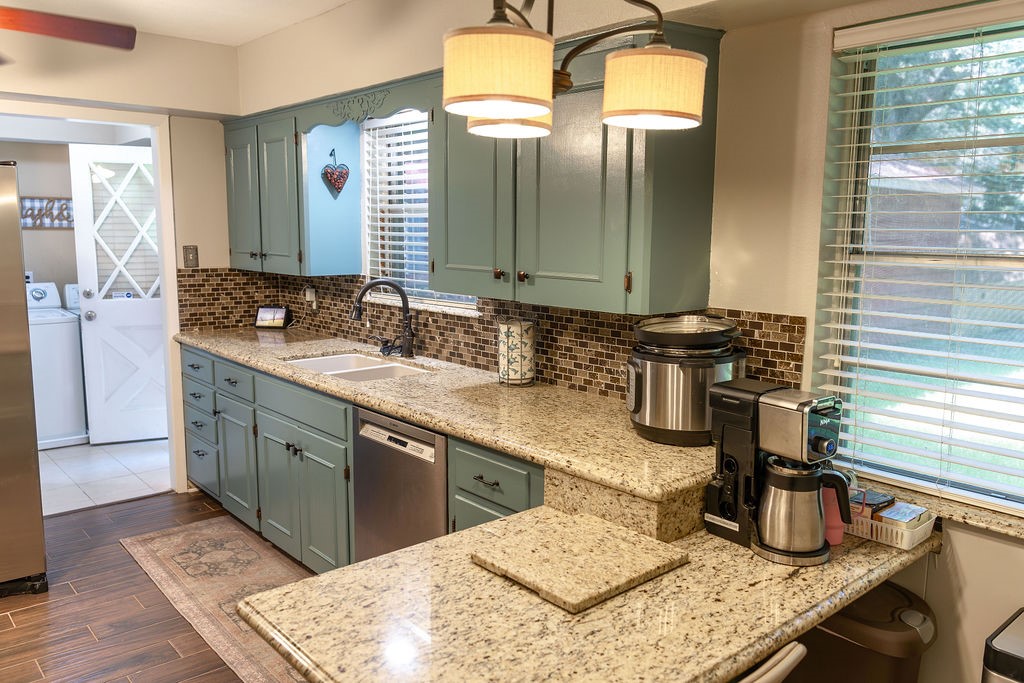 This kitchen features stylish light blue cabinets, granite countertops, and a mosaic tile backsplash. It's equipped with modern appliances and offers ample natural light through a large window. There's a spacious connection to the laundry area.