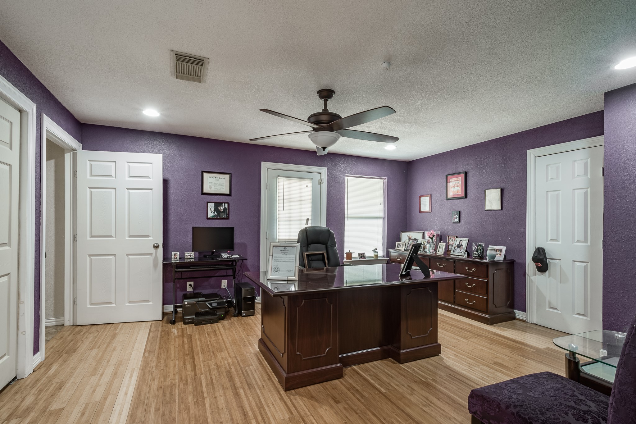 Home office features rich purple walls, elegant wood flooring, and ample natural light. The room is equipped with a large executive desk, shelving for storage, and a ceiling fan for comfort.