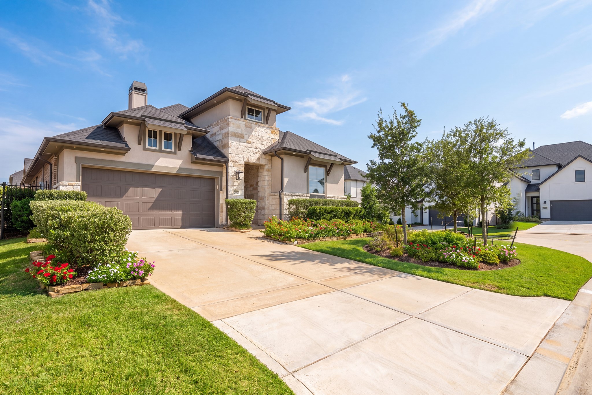 Daytime curb view of the brick and stone facade, extended driveway and two-car garage, framed by tidy shrubs, young shade trees and a crisp, well-maintained front lawn.
