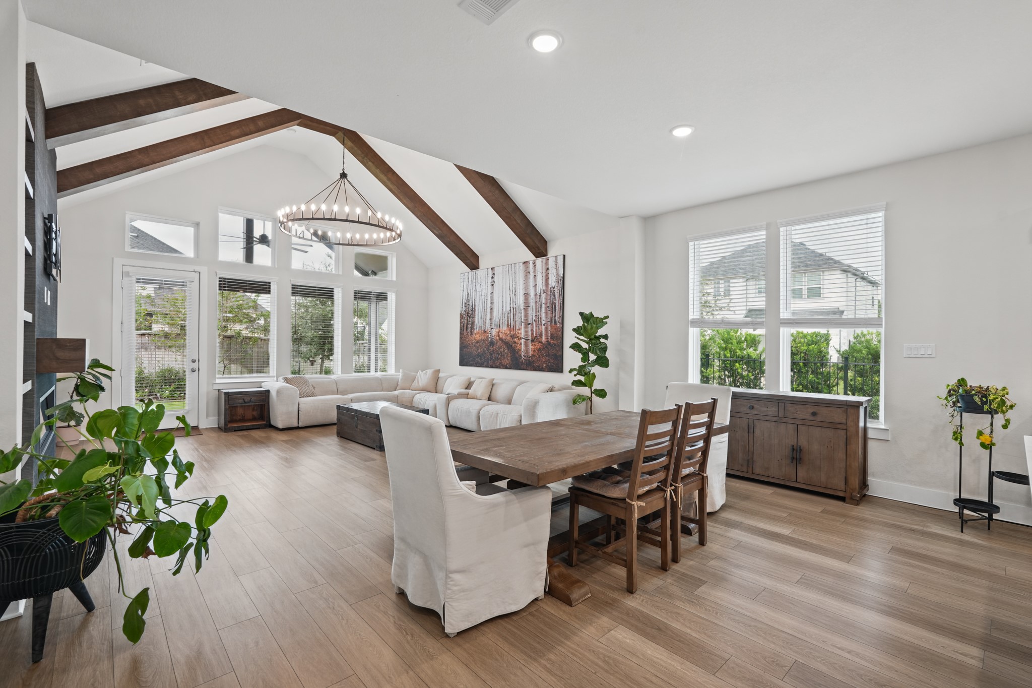 The dining space sits beneath the vaulted, beamed ceiling with windows on two sides, flowing directly into both the kitchen and the living room in one continuous open area.