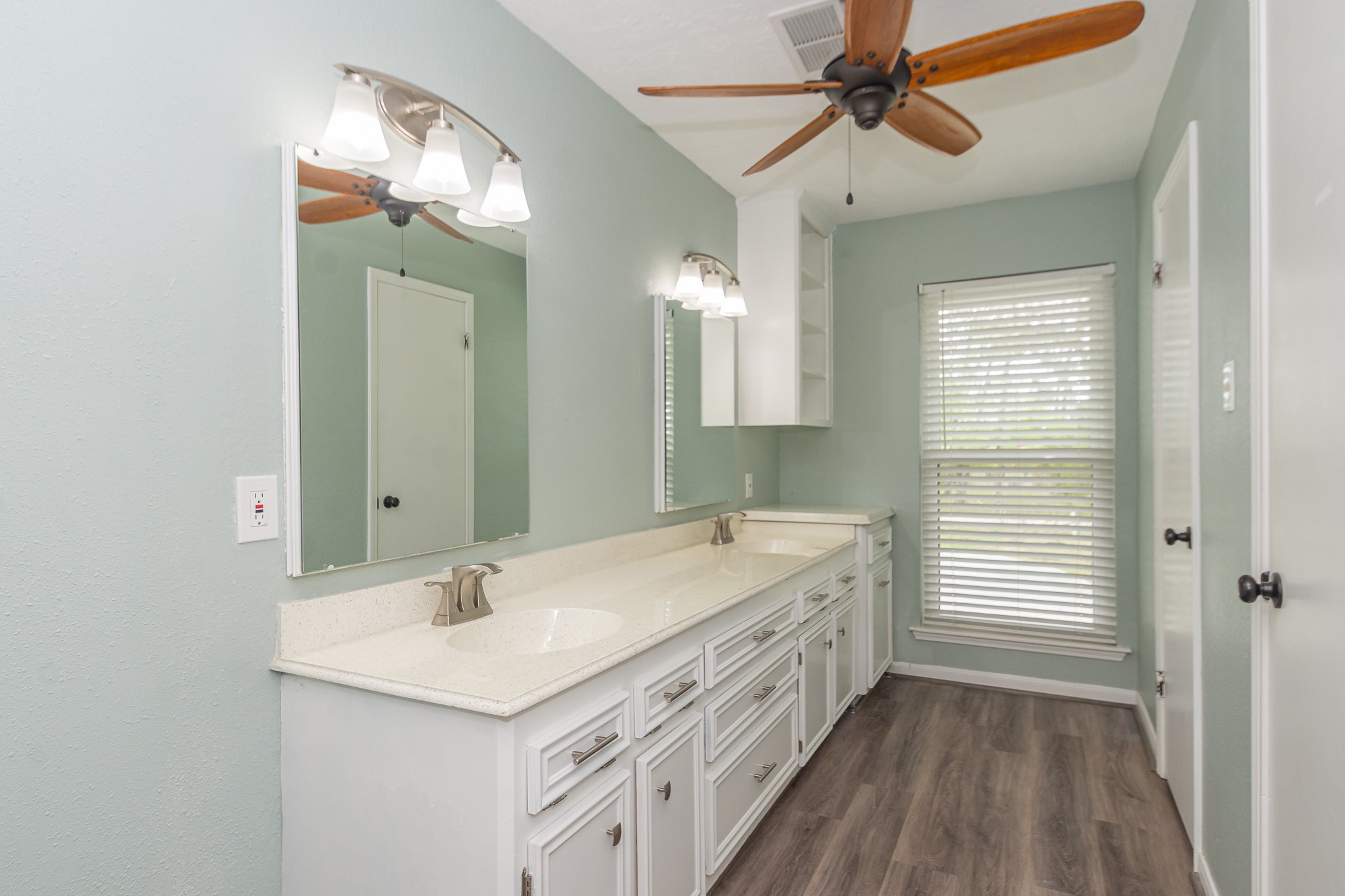 Primary bathroom with a long dual-sink vanity and bright lighting. One door leading to the toilet and stand-up shower, the other into a walk-in closet.