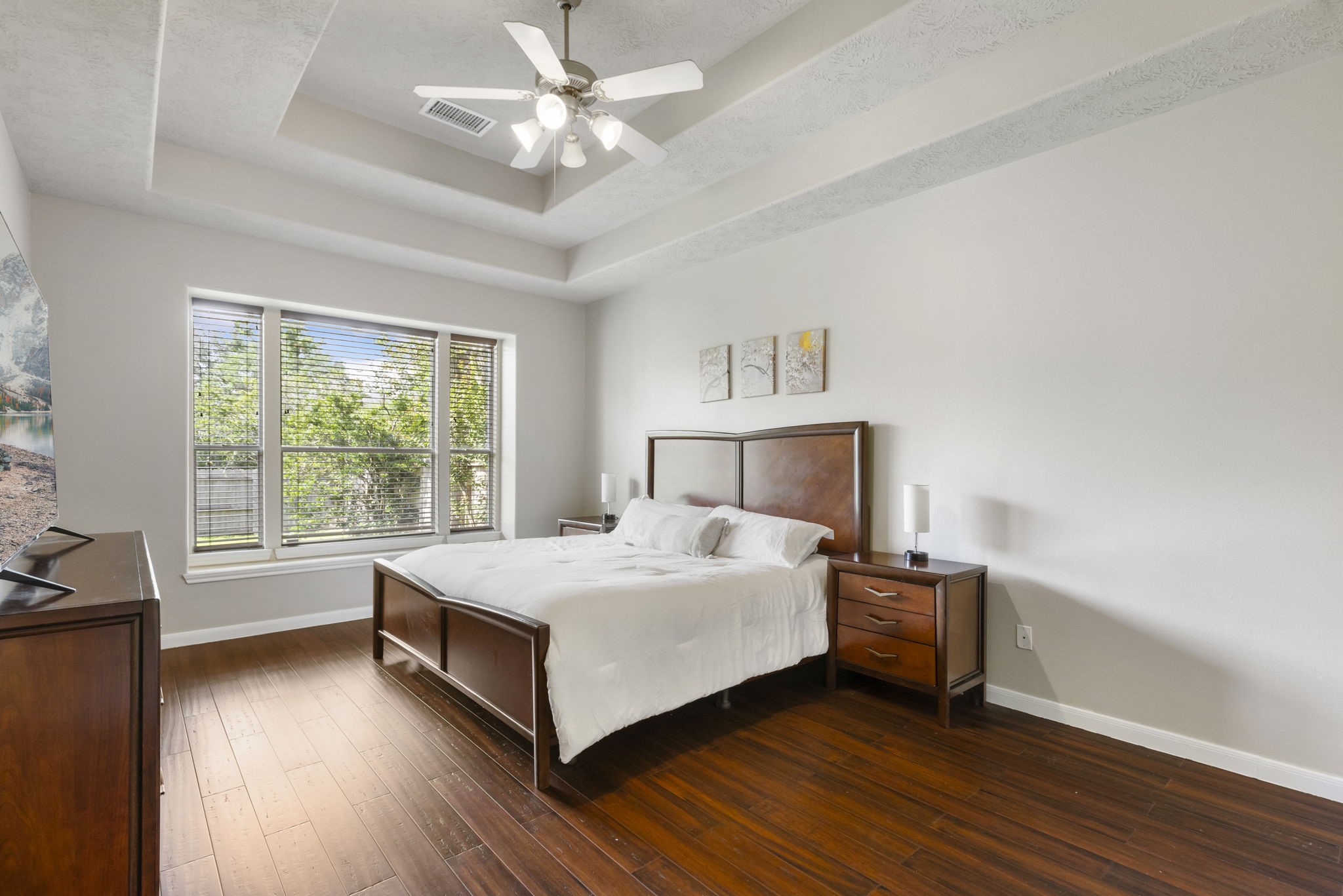 The first-floor primary suite offers a peaceful retreat featuring beautiful wood flooring, a tray ceiling, and large windows overlooking the backyard greenery.