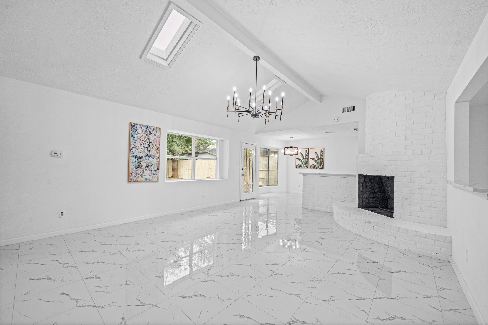 Bright and spacious living area featuring a vaulted ceiling with skylight, elegant chandelier, and a cozy white brick fireplace, complemented by glossy marble-style flooring and large windows for abundant natural light.
