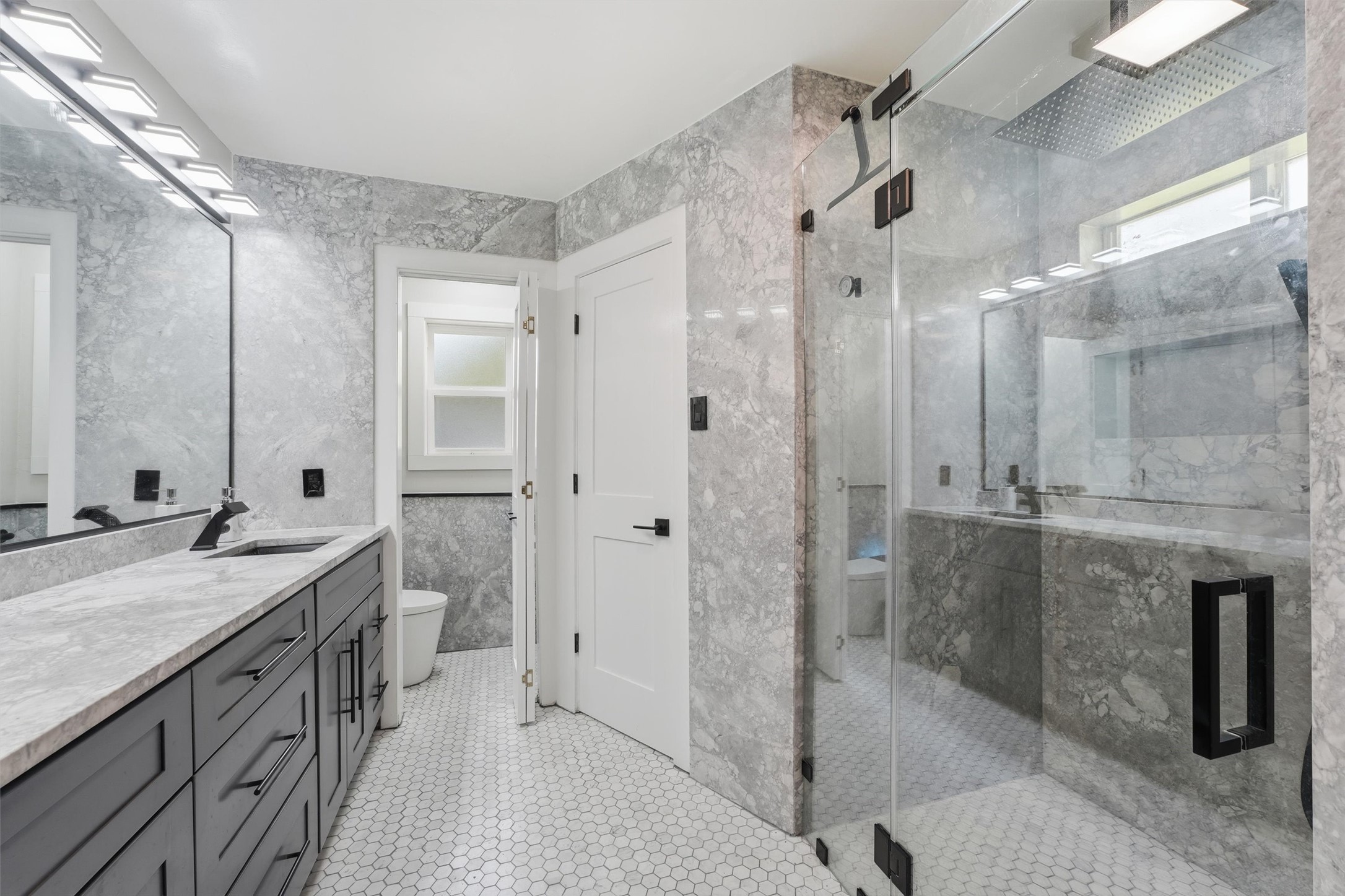 The main primary bathroom features a large glass-enclosed walk-in shower with natural quartzite walls, a double vanity with black fixtures, distinctive hexagonal tile flooring, and closets on either side of the shower.