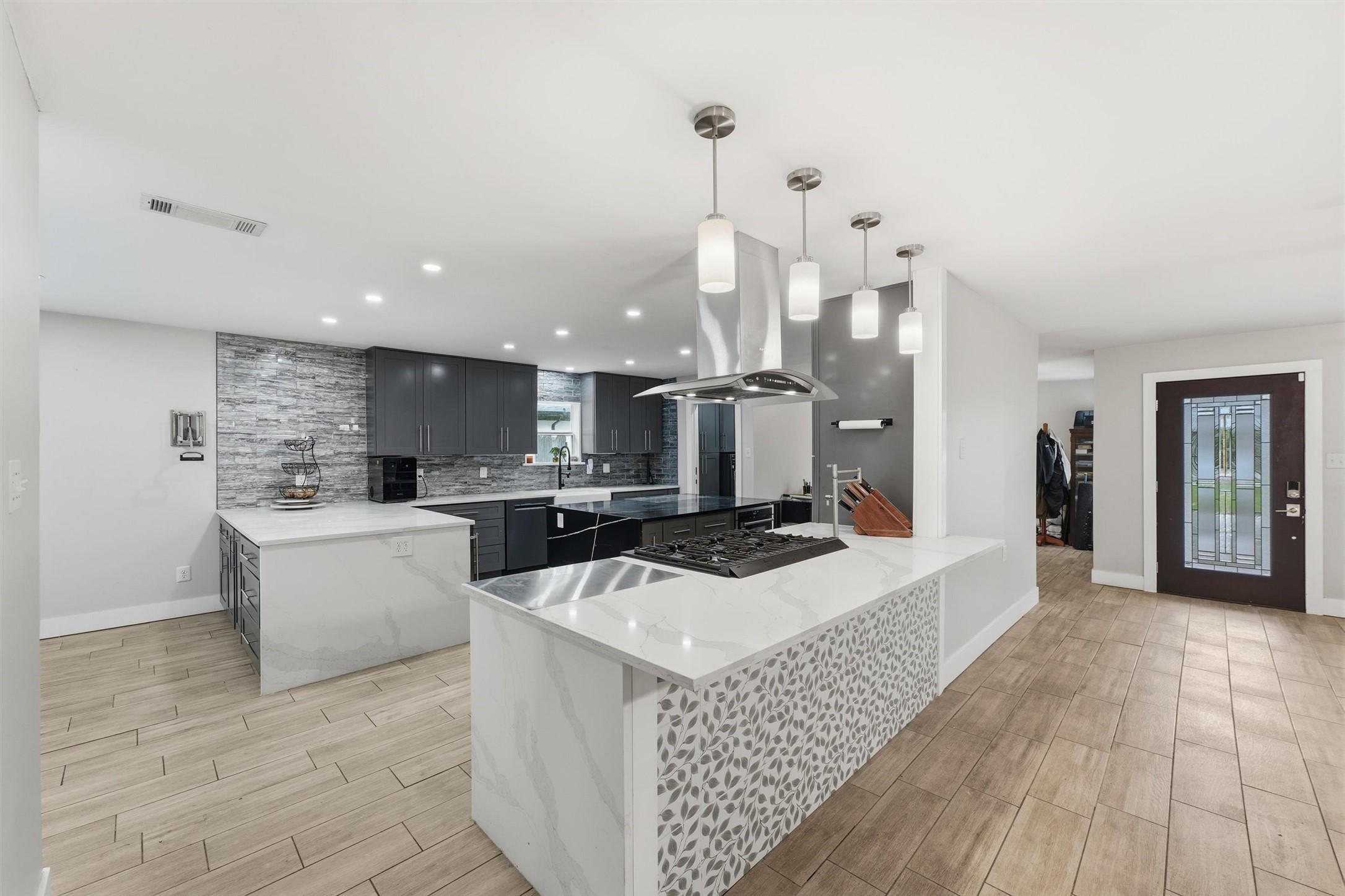 The kitchen offers extensive counterspace, dark cabinetry, a gas cooktop with a convenient pot filler, stainless steel range hood, pendant lighting, and decorative tile accents.