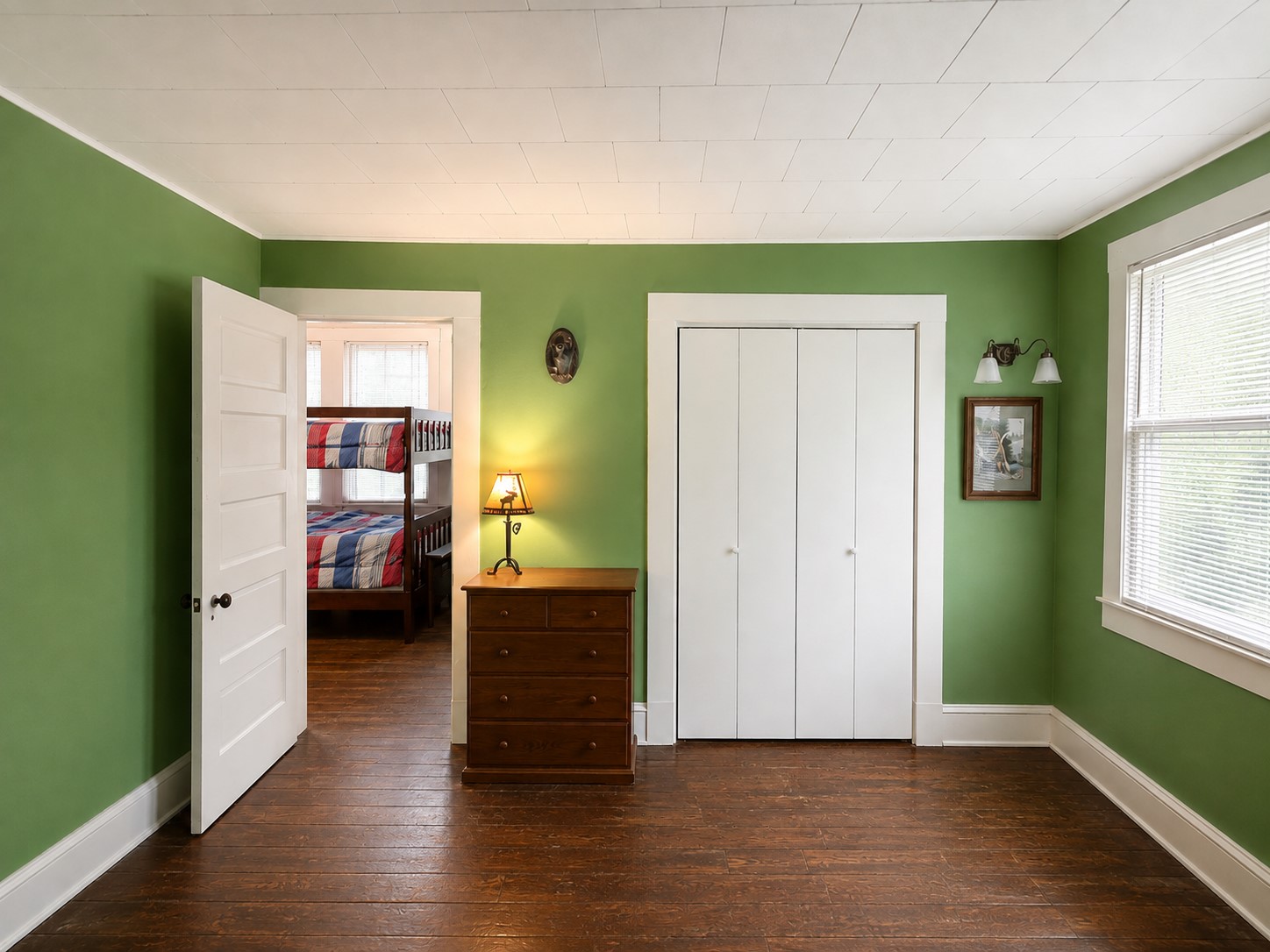 Spacious upstairs bedroom features rich wood flooring, green walls, classic white trim, and a large closet. A wide window brings natural light into the room, while the open floor area allows for flexible furniture placement.