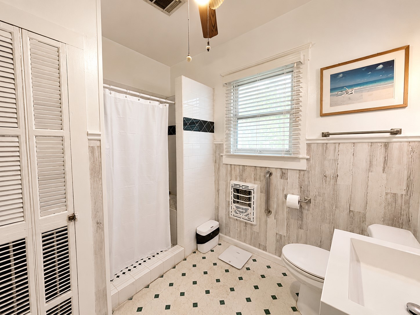 Bright bathroom features a tiled shower with contrasting accent detail, abundant natural light, and classic trim. Wood-look wall treatment adds texture while complementing the rooms light, neutral palette.
