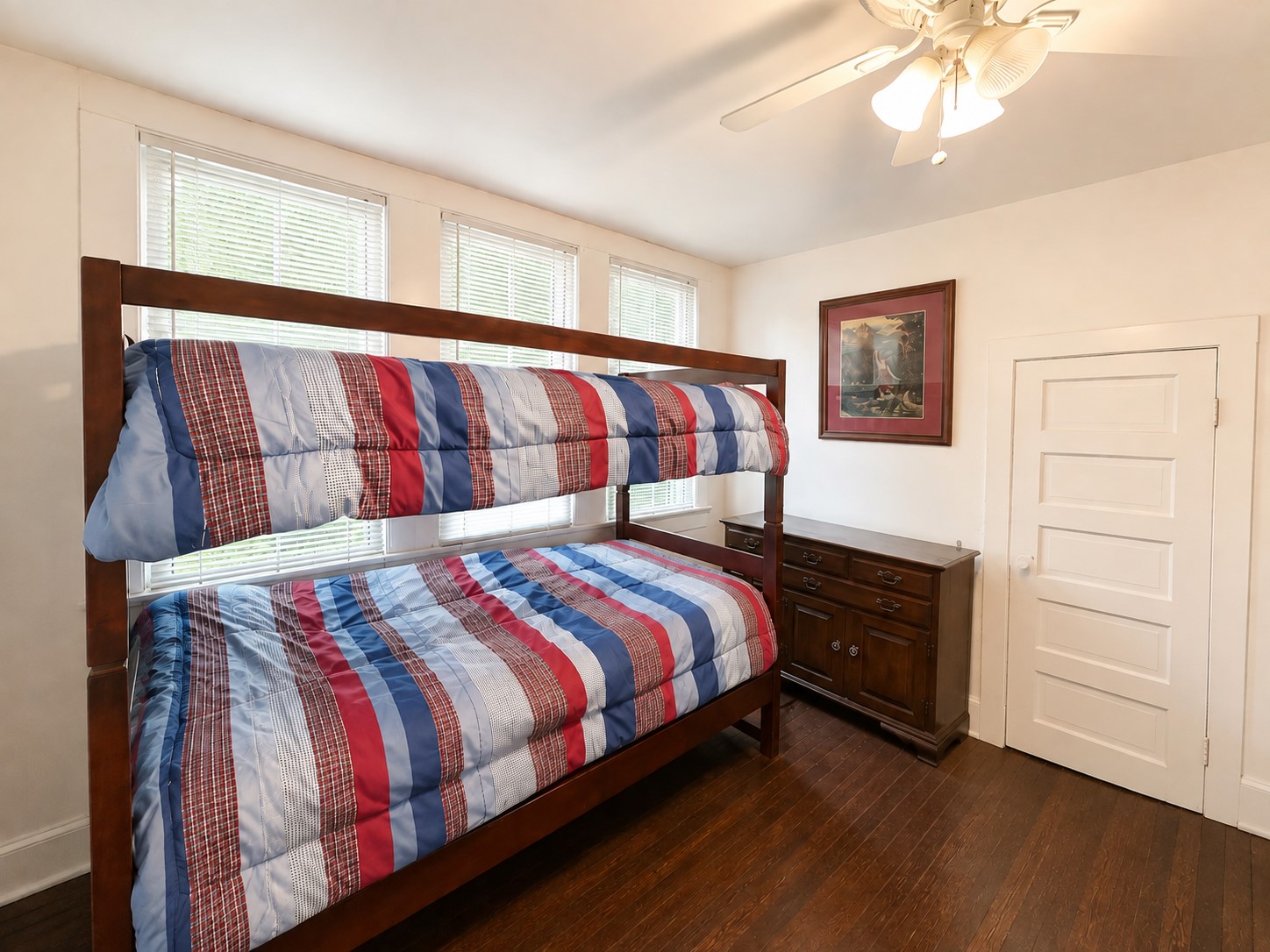 A second angle highlights the rooms generous window arrangement, wood flooring, and ample space for furnishings. The ceiling fan adds everyday functionality.
