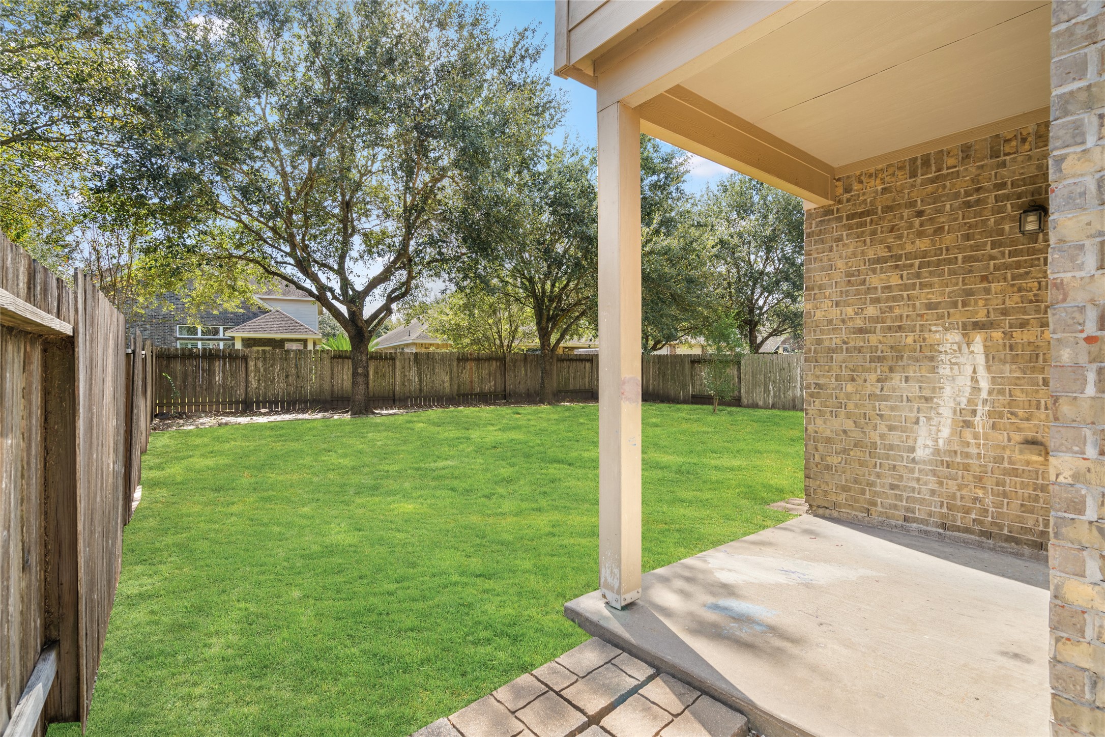Covered backyard patio overlooking a green lawn and mature trees. The shaded outdoor area provides a comfortable spot for outdoor dining, relaxing, or enjoying the backyard in warmer weather.