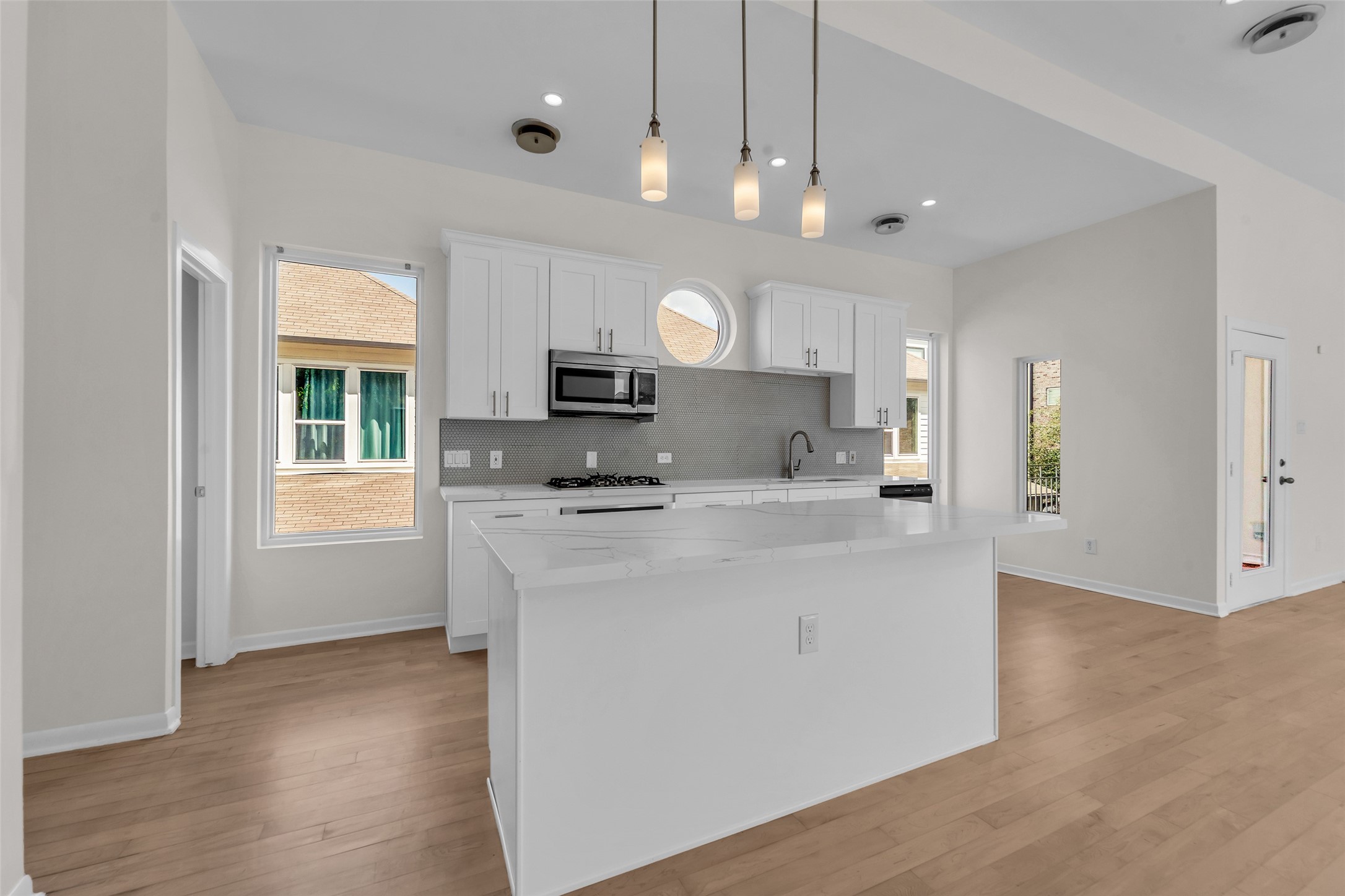 Bright and modern kitchen featuring white cabinetry, stainless steel appliances, and a spacious island with pendant lighting, all set against warm wood flooring.
